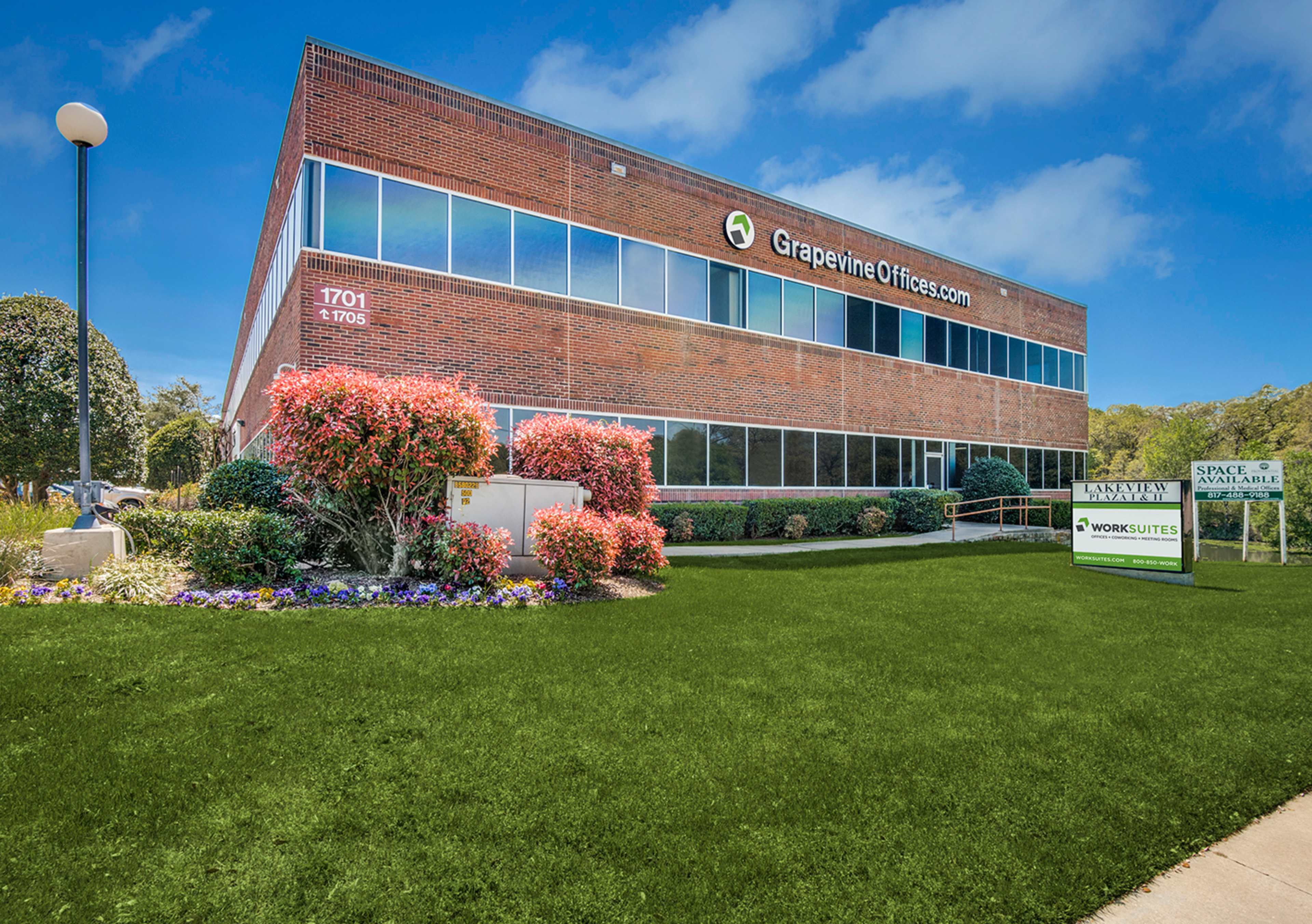 A brick office building with large windows is set in a landscaped area featuring colorful flowers and a sign indicating available space for rent.