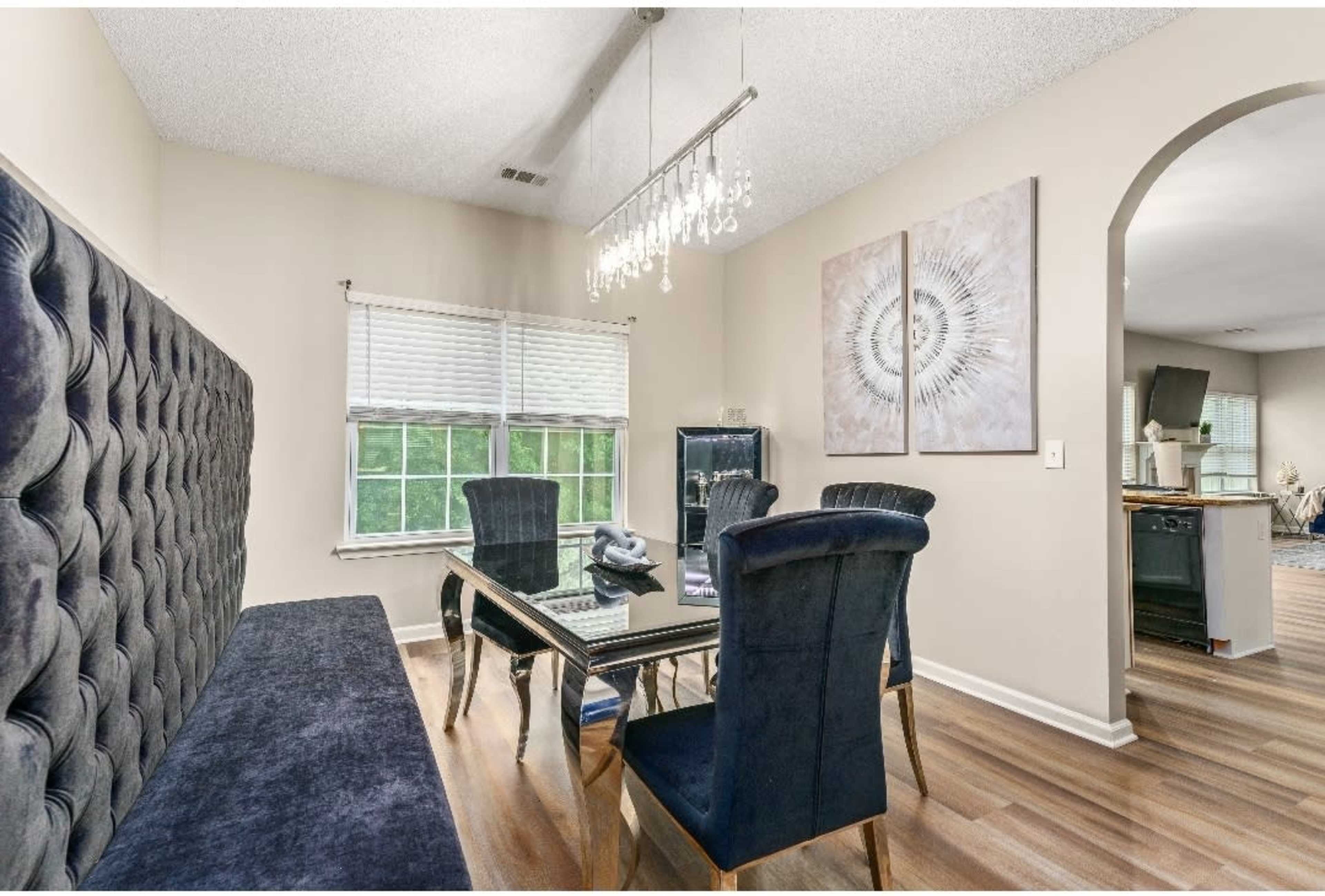 The dining area features a sleek black table surrounded by four upholstered chairs, alongside a tufted bench and a decorative chandelier.