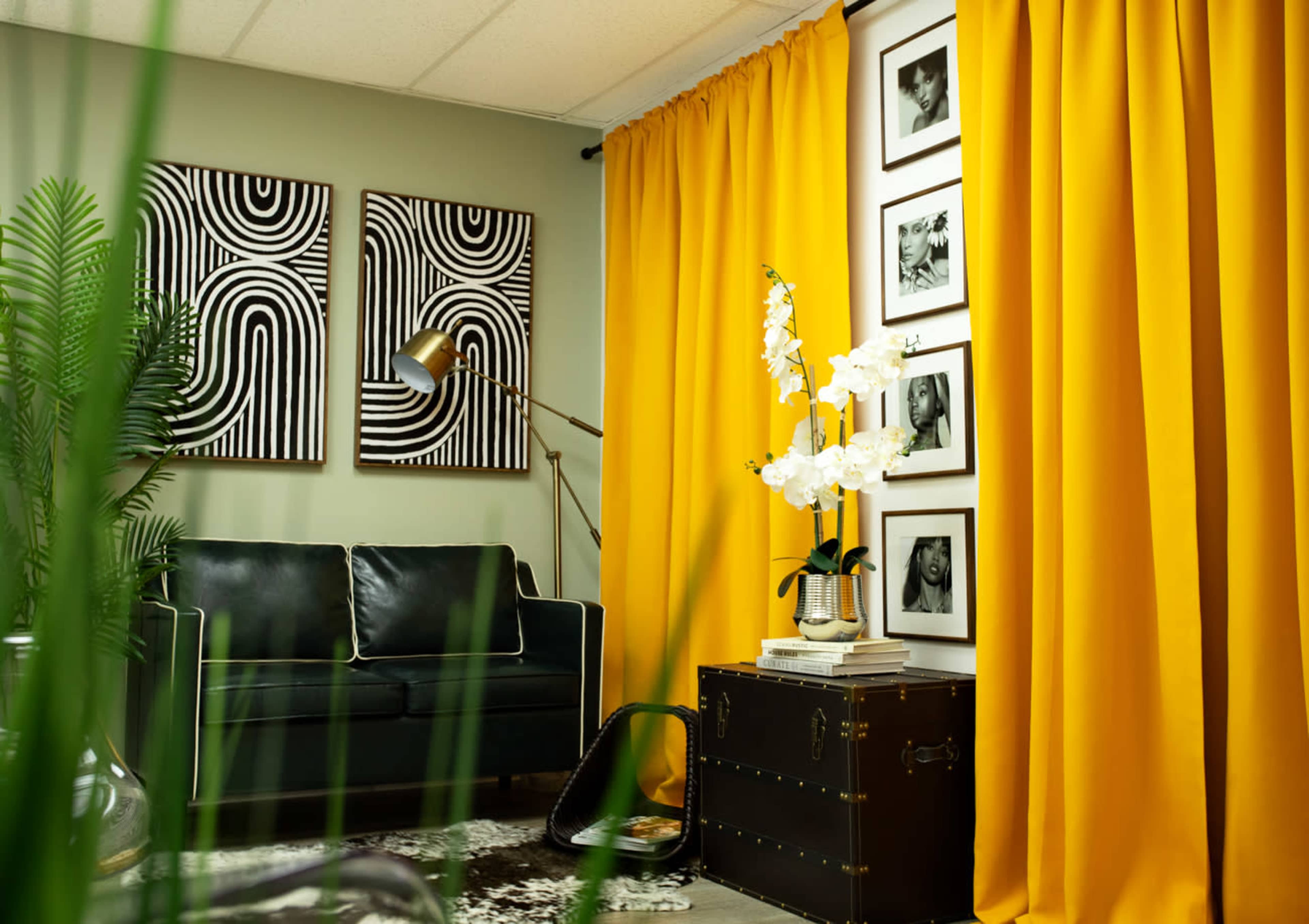 The image features a modern interior with a black couch, yellow curtains, a decorative lamp, framed black-and-white art, and a coffee table topped with white orchids.