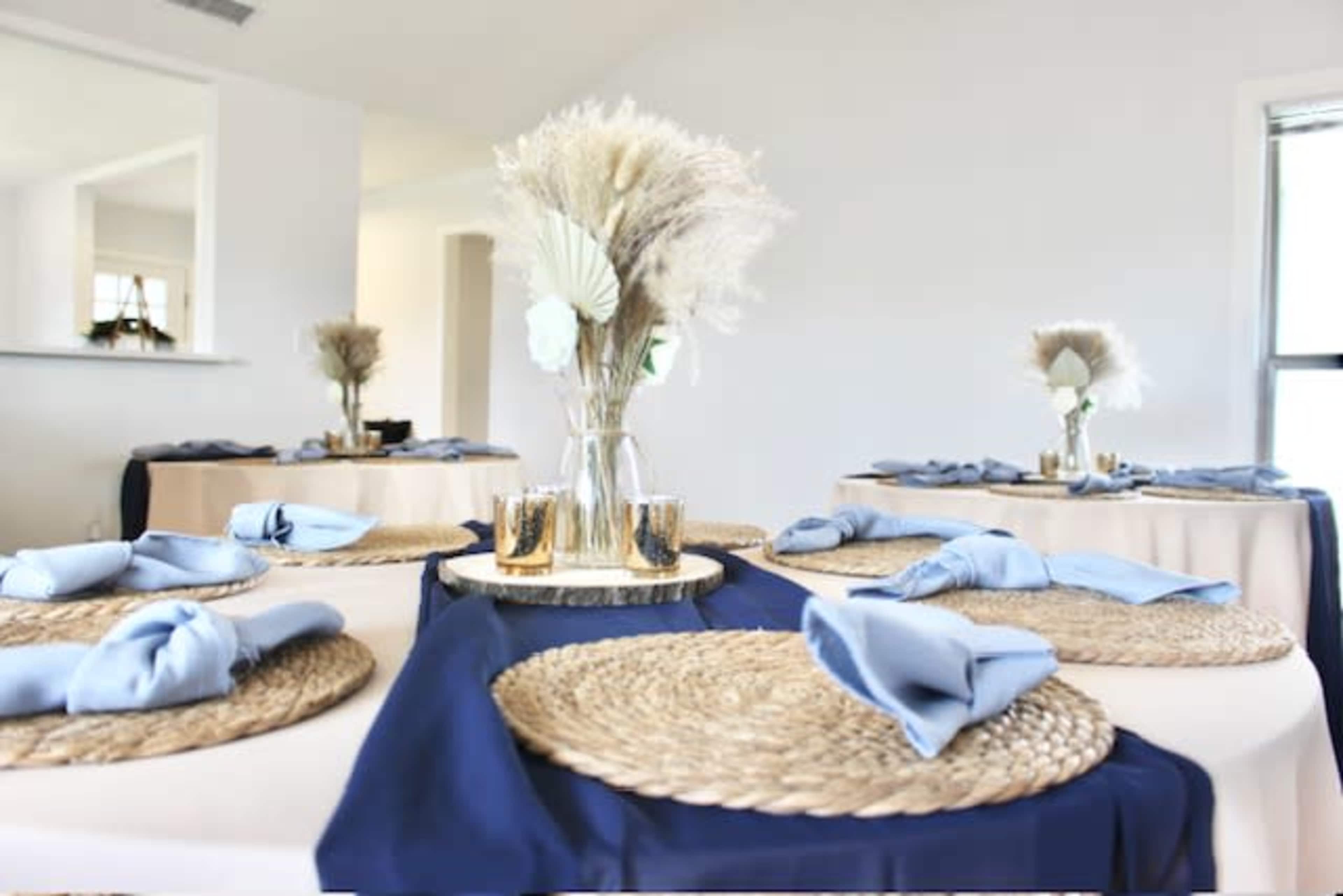 A dining area features round tables adorned with blue and beige tablecloths, woven placemats, and vases of dried flowers.