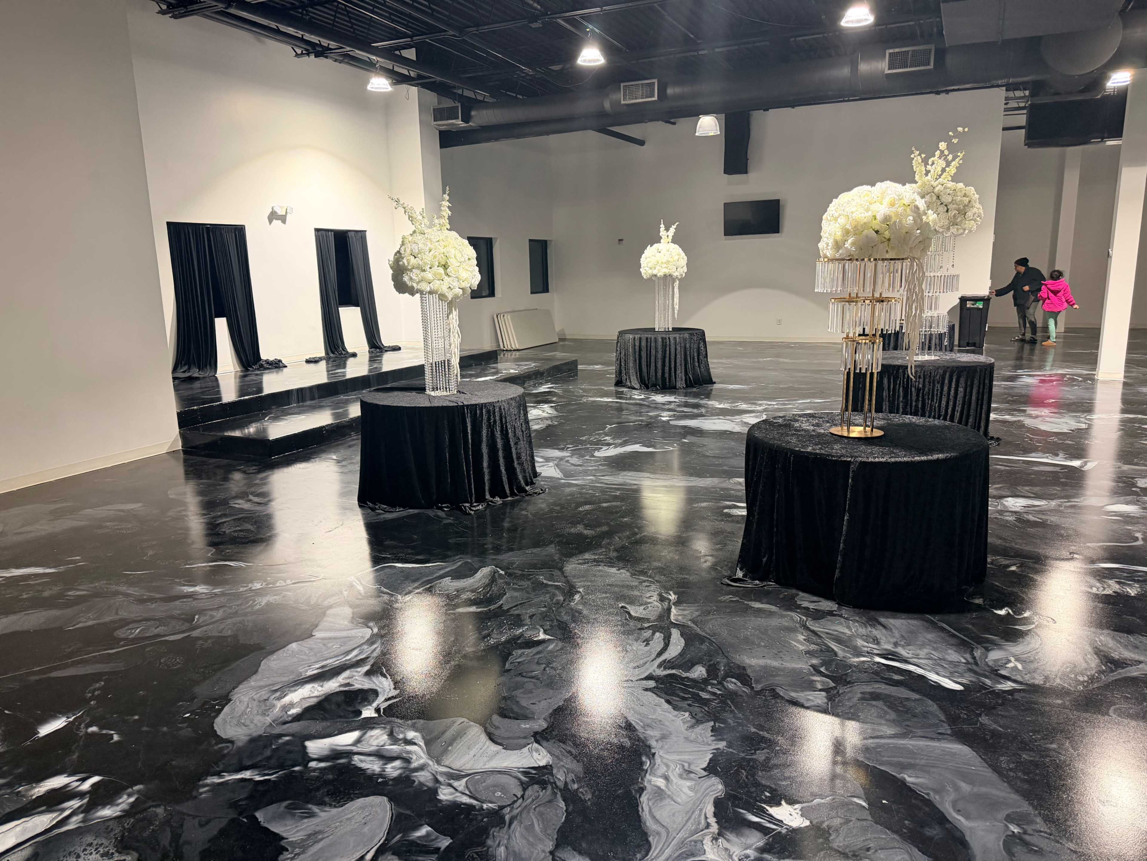 An event space features four round tables with black tablecloths and floral centerpieces, arranged on a glossy black and white marbled floor.