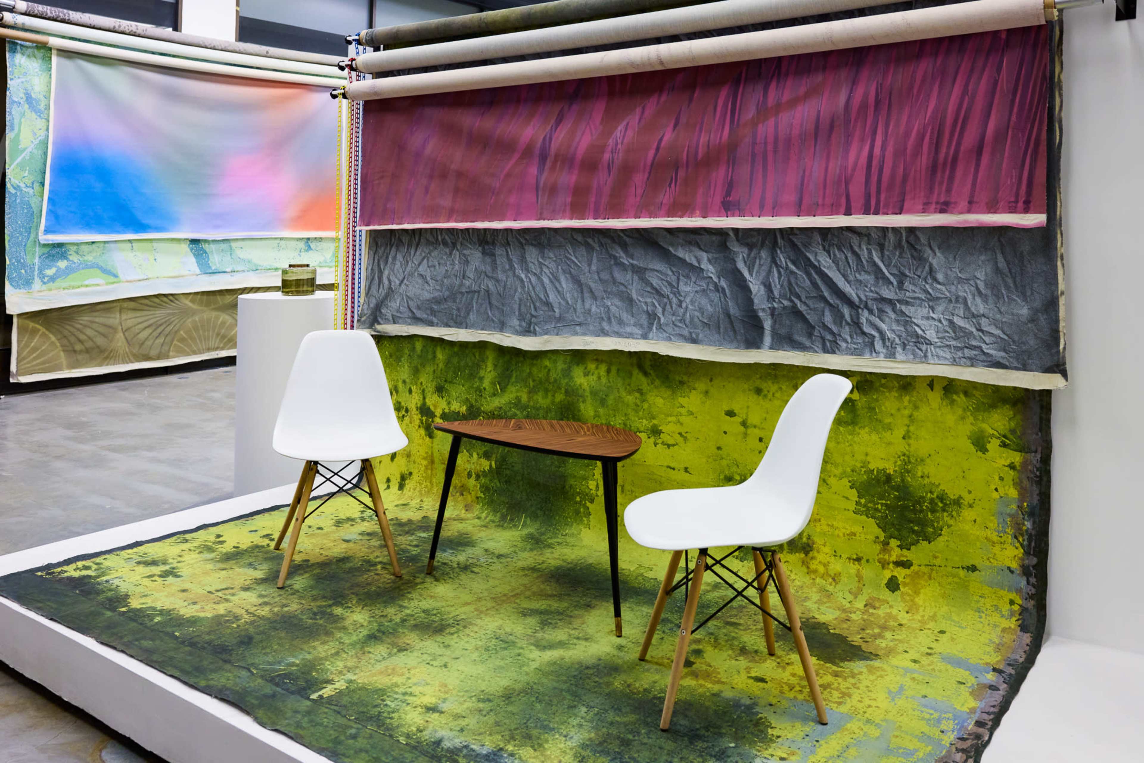The image shows a minimalist art installation featuring two white chairs and a small wooden table on a colorful, textured backdrop of fabric.