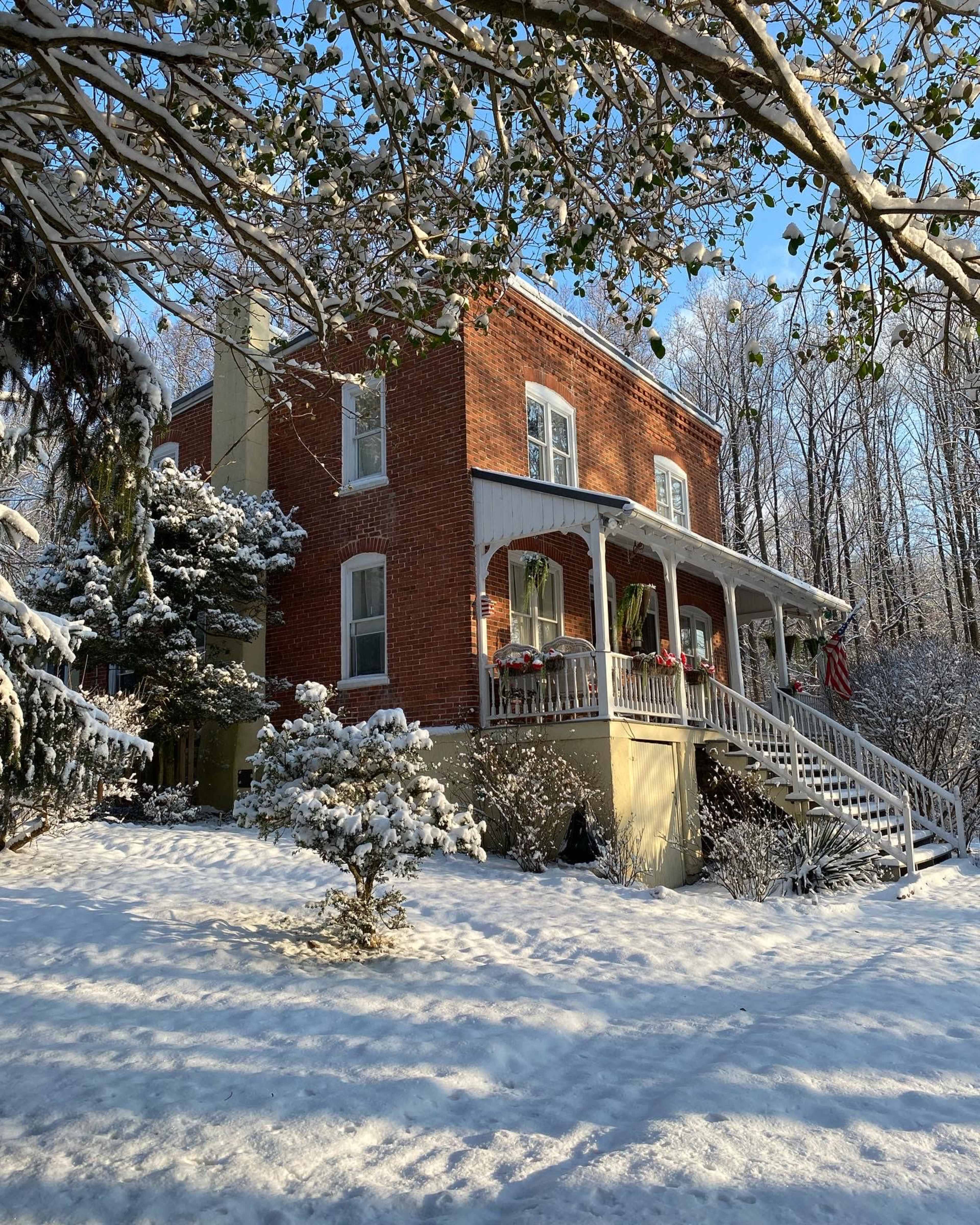 A two-story brick house with a front porch is surrounded by snow-covered trees and bushes on a sunny winter day.