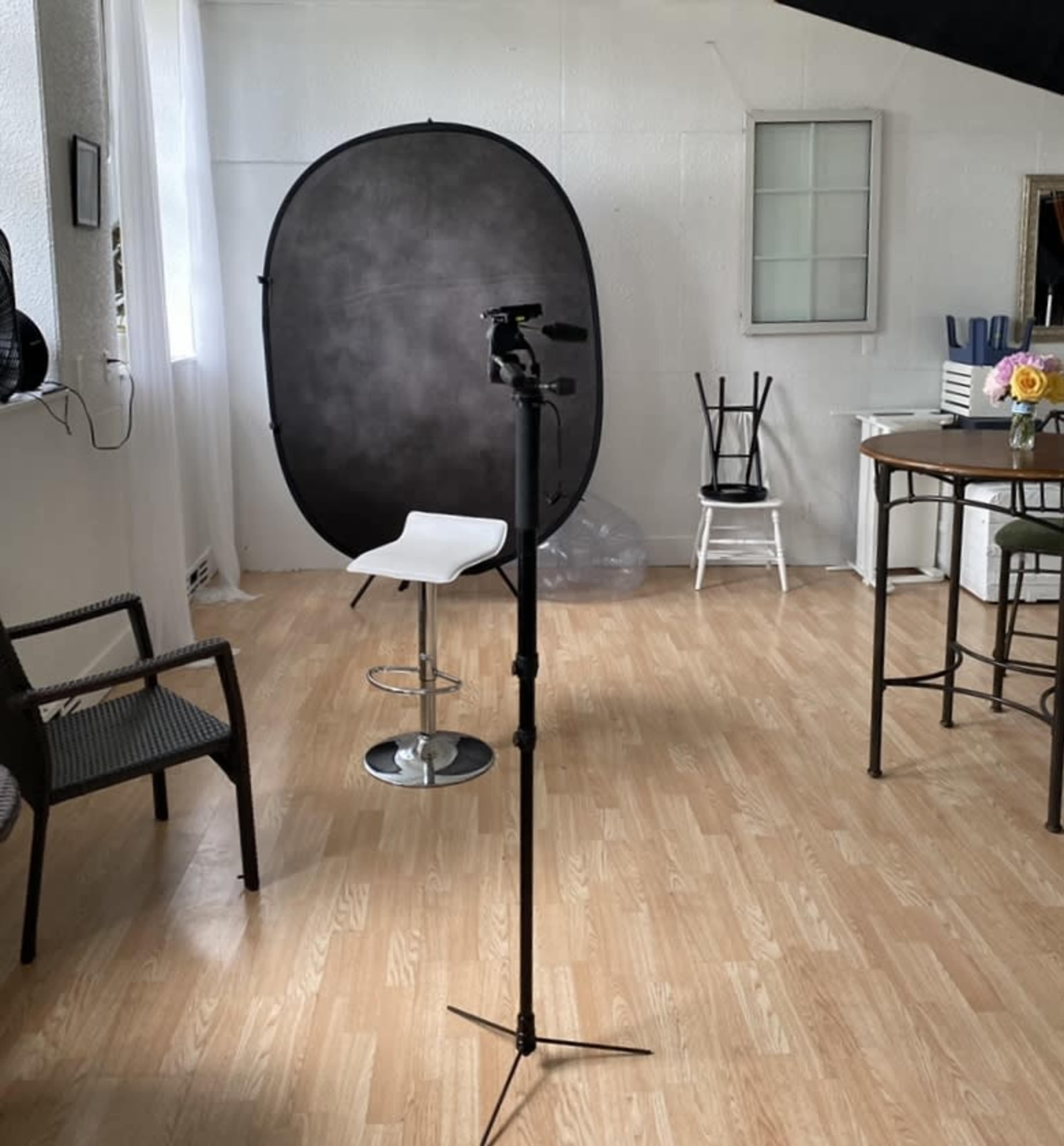 A photography studio setup featuring a backdrop, a camera on a tripod, and various chairs around a wooden table.