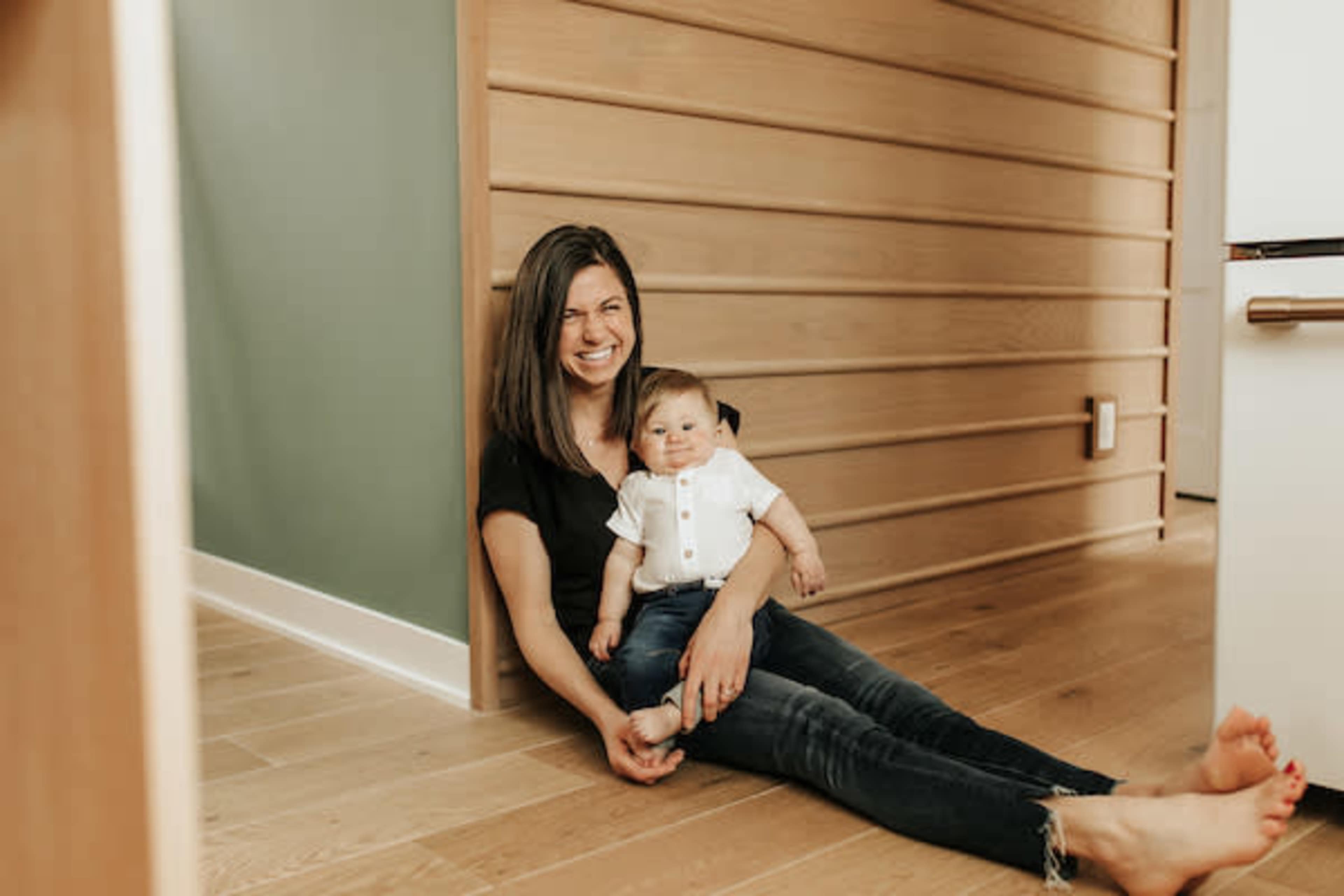 A woman sits on the floor against a wall, holding an infant who is smiling in her lap.