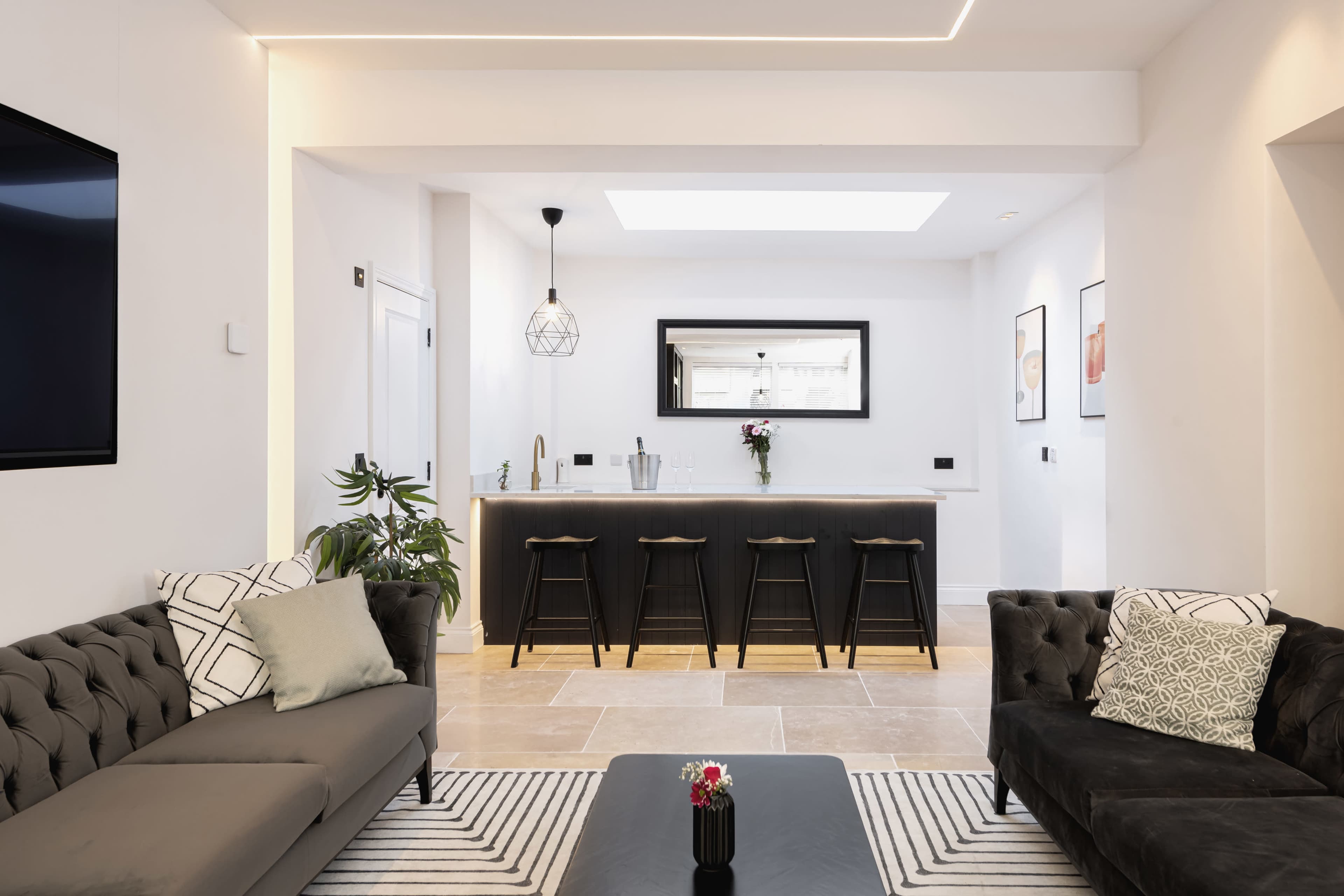A modern living space features a sitting area with dark sofas, a coffee table, and a kitchen bar with high stools in the background.