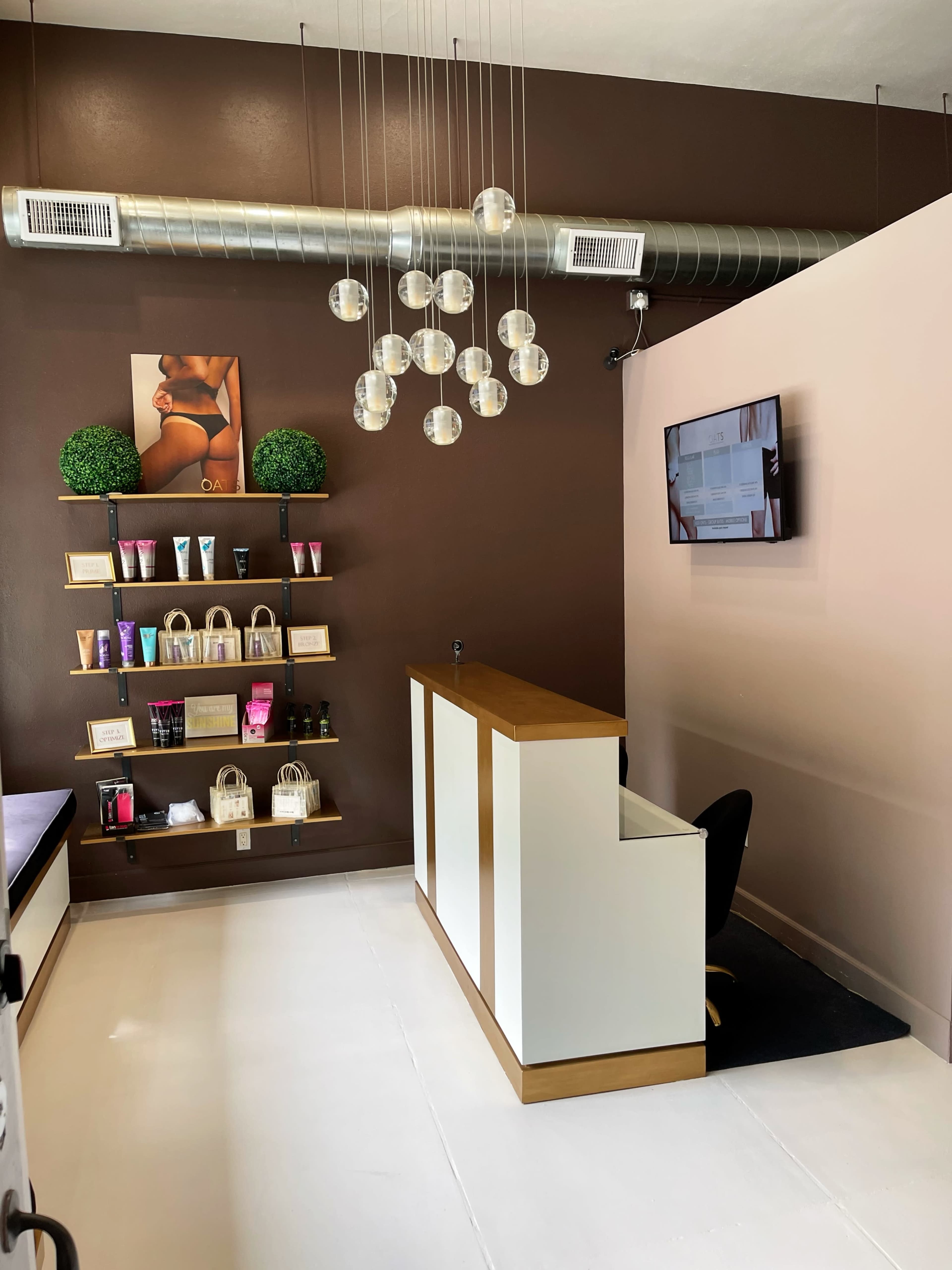 The image shows a modern reception area with a sleek front desk, a wall-mounted television, decorative plants on shelves, and a variety of beauty products displayed.