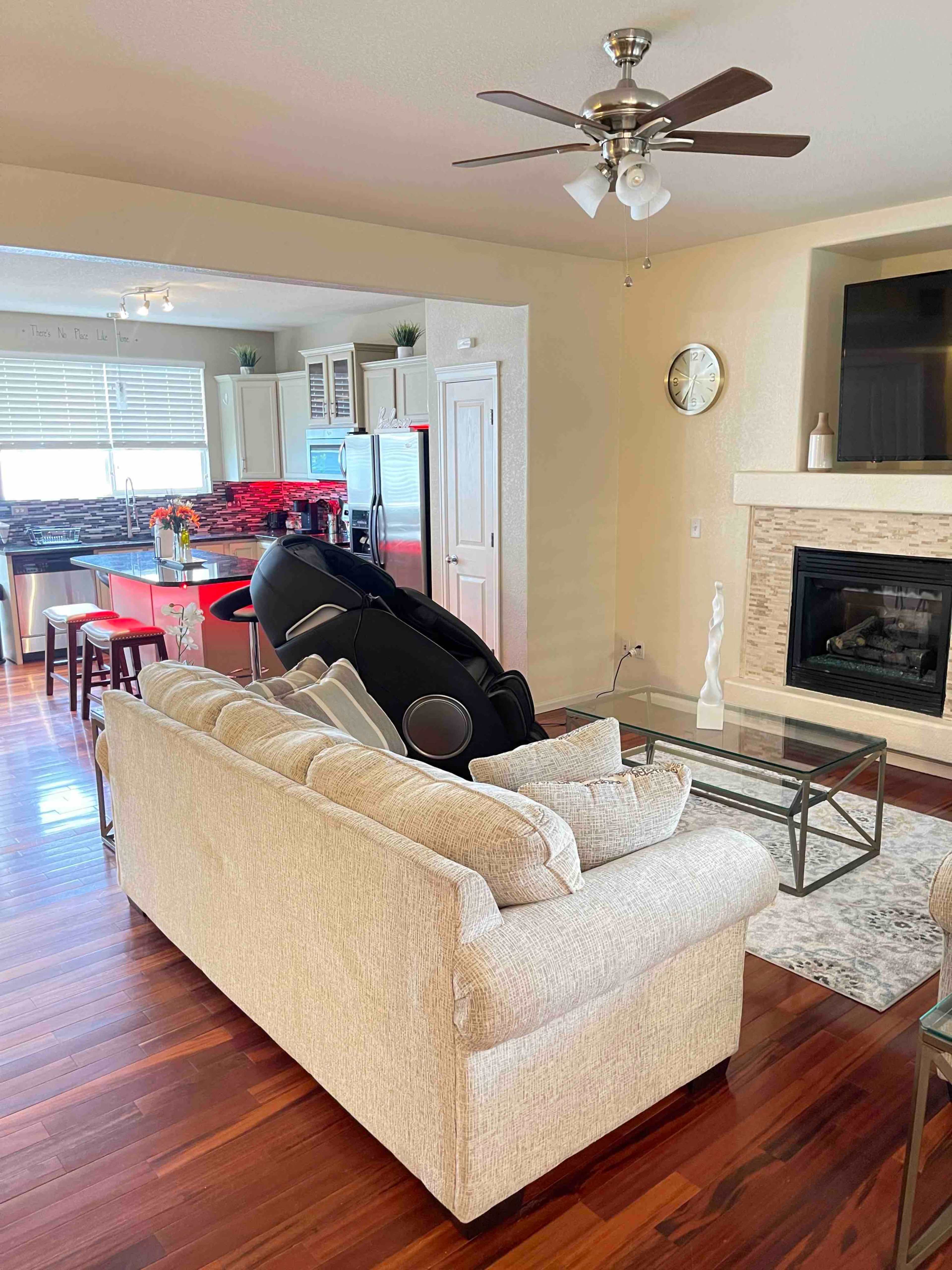 A well-lit living room features a beige sofa, a glass coffee table, and a modern kitchen in the background with red accents and bar seating.