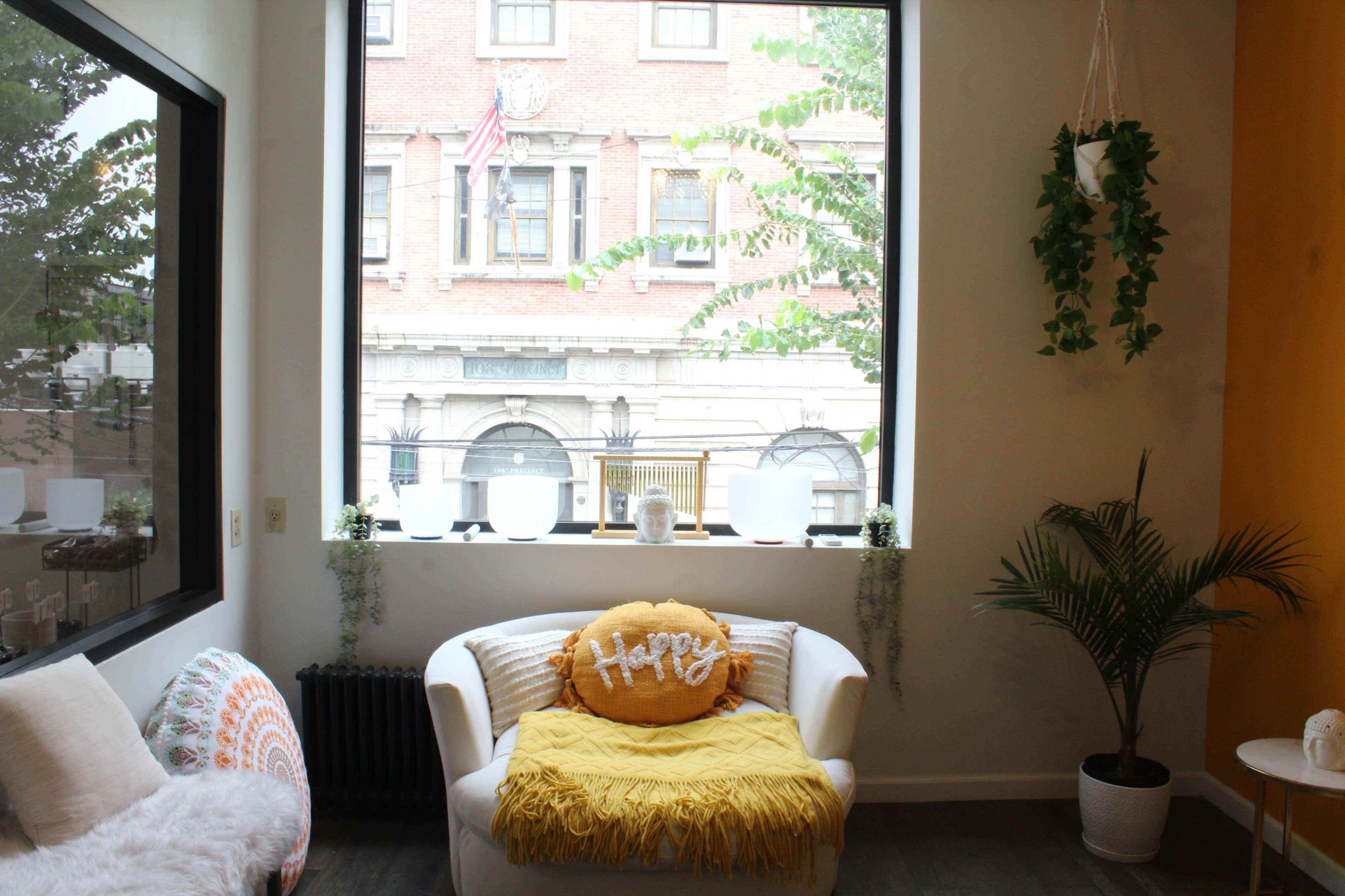 A cozy living space features a white chair with a yellow throw pillow and a window showcasing a brick building with flags.