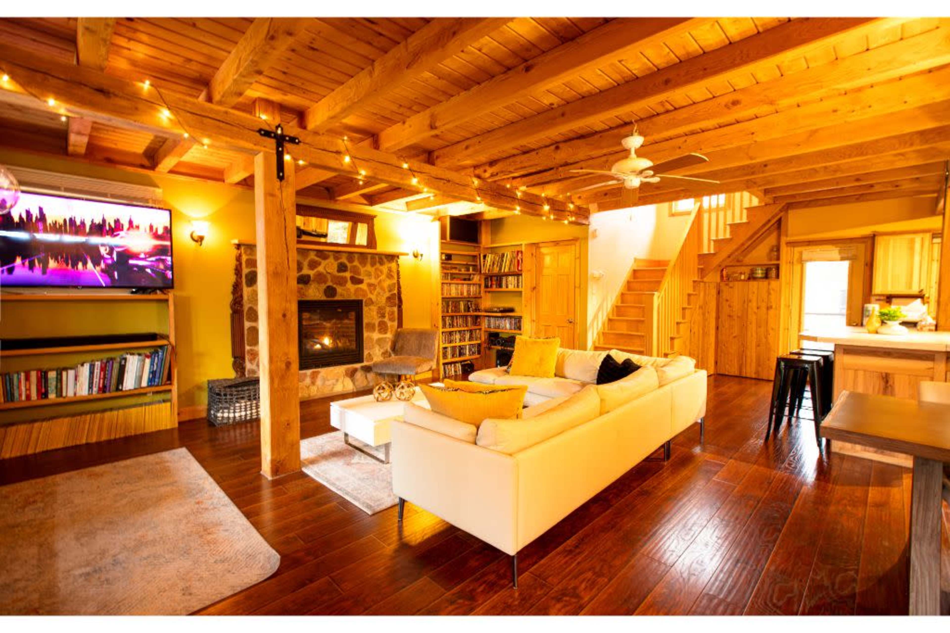The image shows a spacious, wooden-floored living room with a white sofa, a stone fireplace, a wall-mounted television, and a staircase leading to an upper level.