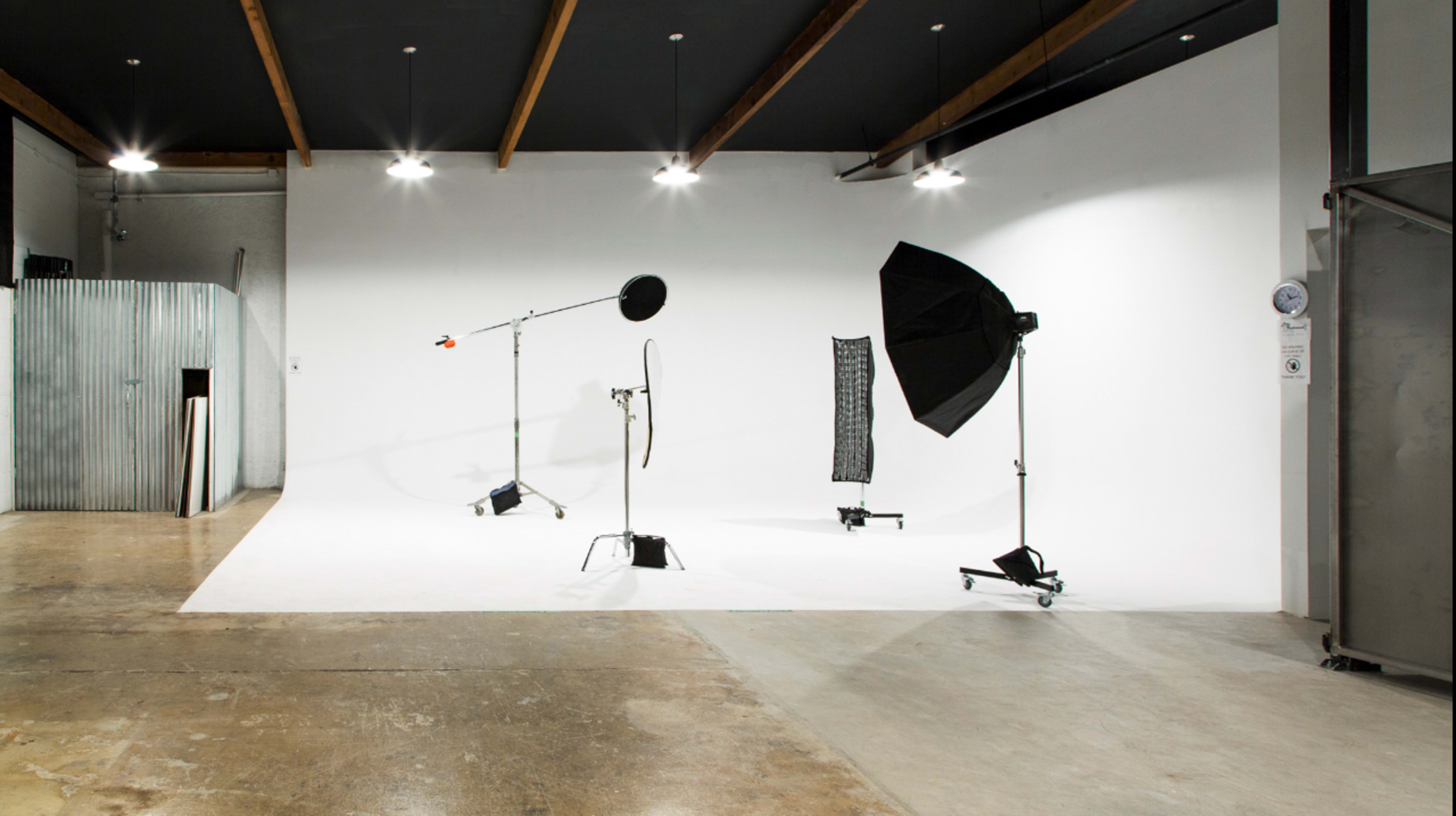 The image shows a photography studio with a white backdrop and various lighting equipment, including a softbox and a light stand.