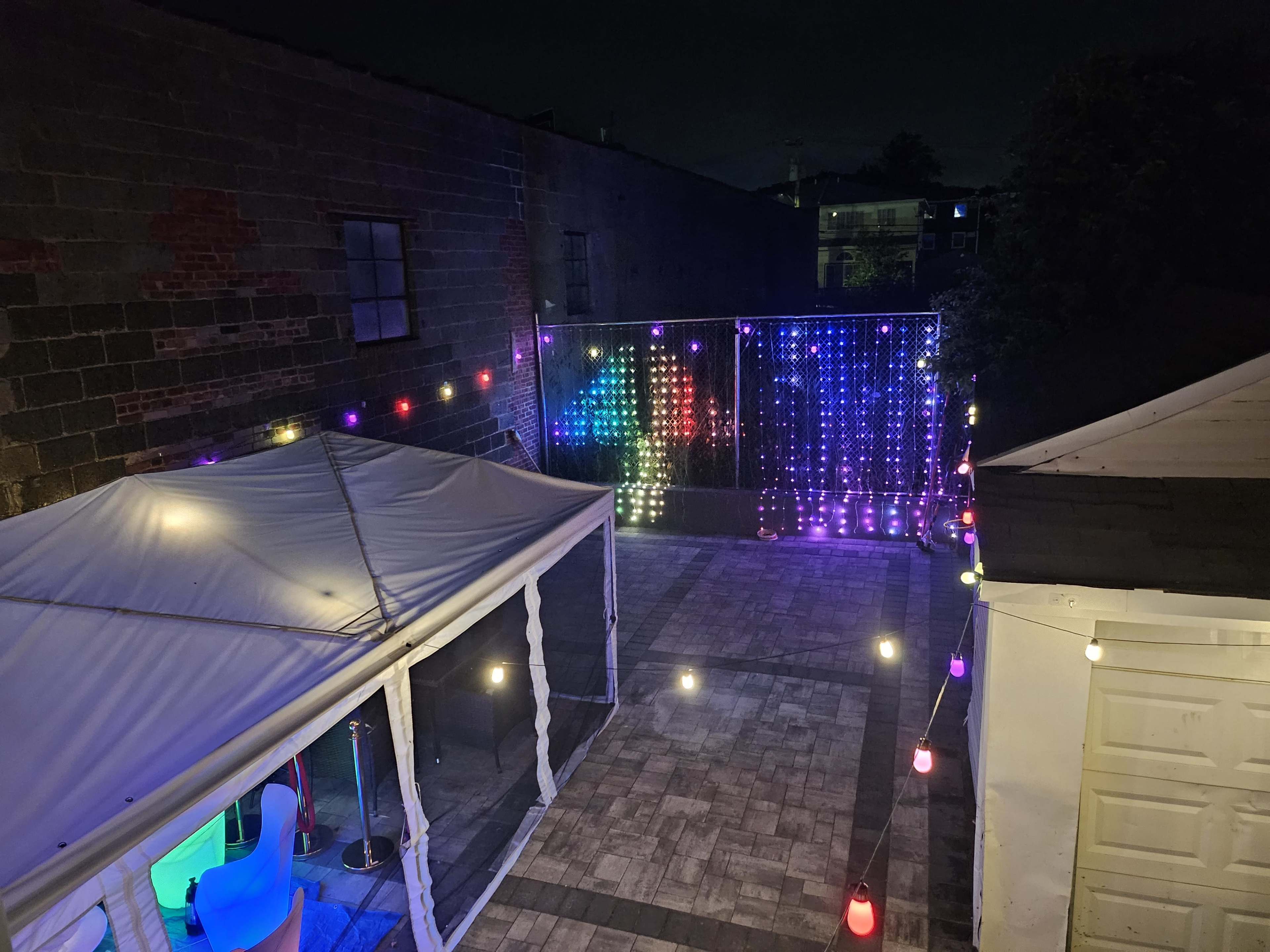 The image shows a spacious outdoor area at night, illuminated by colorful lights and a decorated wall of illuminated strings.