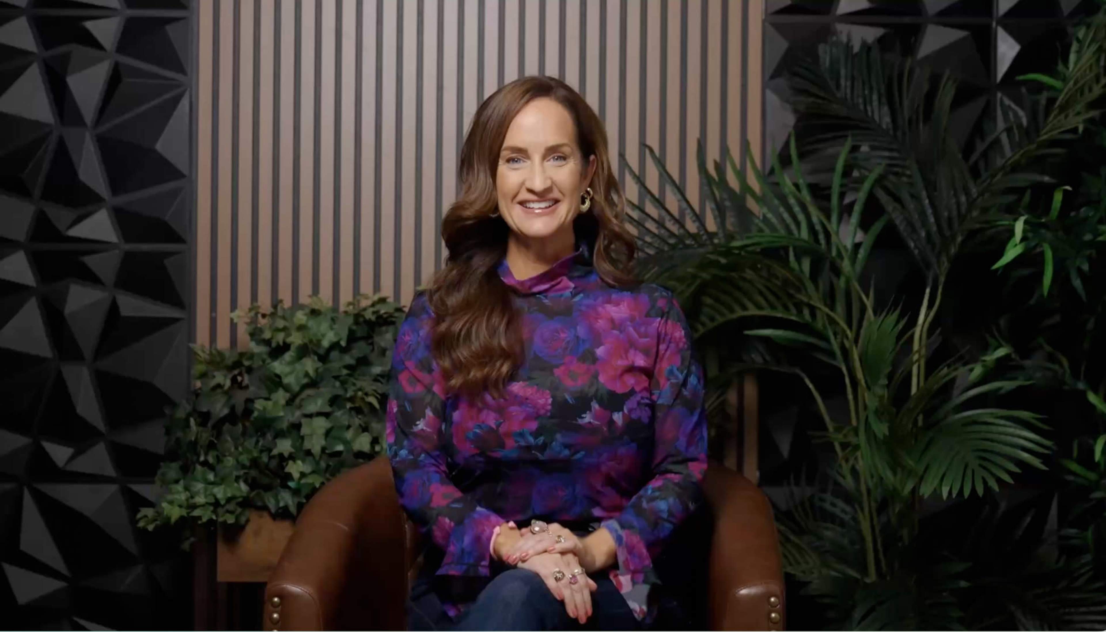 A woman with long hair sits on a brown chair in front of a striped wall and green plants.