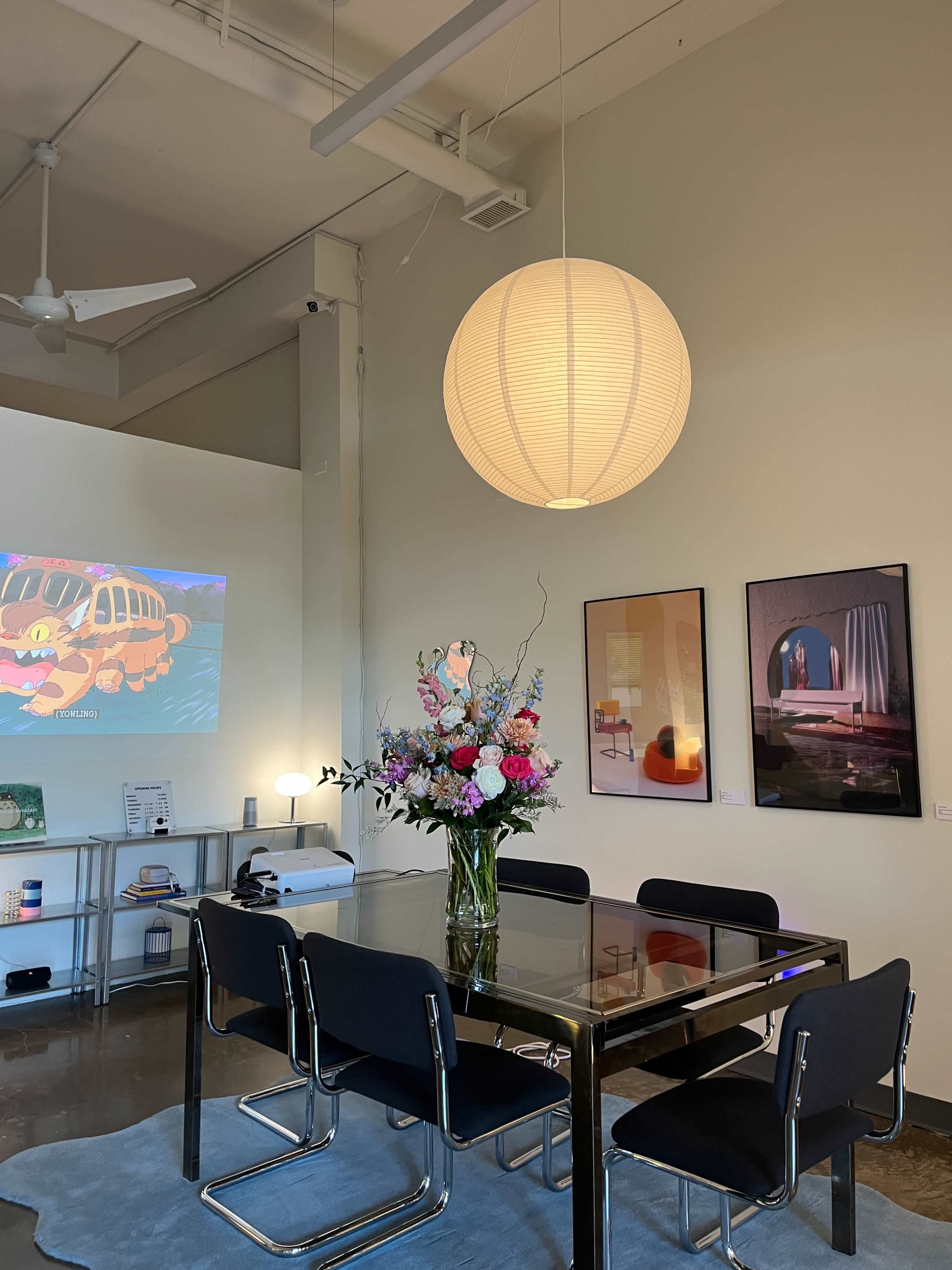 A modern dining area features a glass table surrounded by black chairs, illuminated by a large paper lantern, with floral arrangements and artwork on the walls.