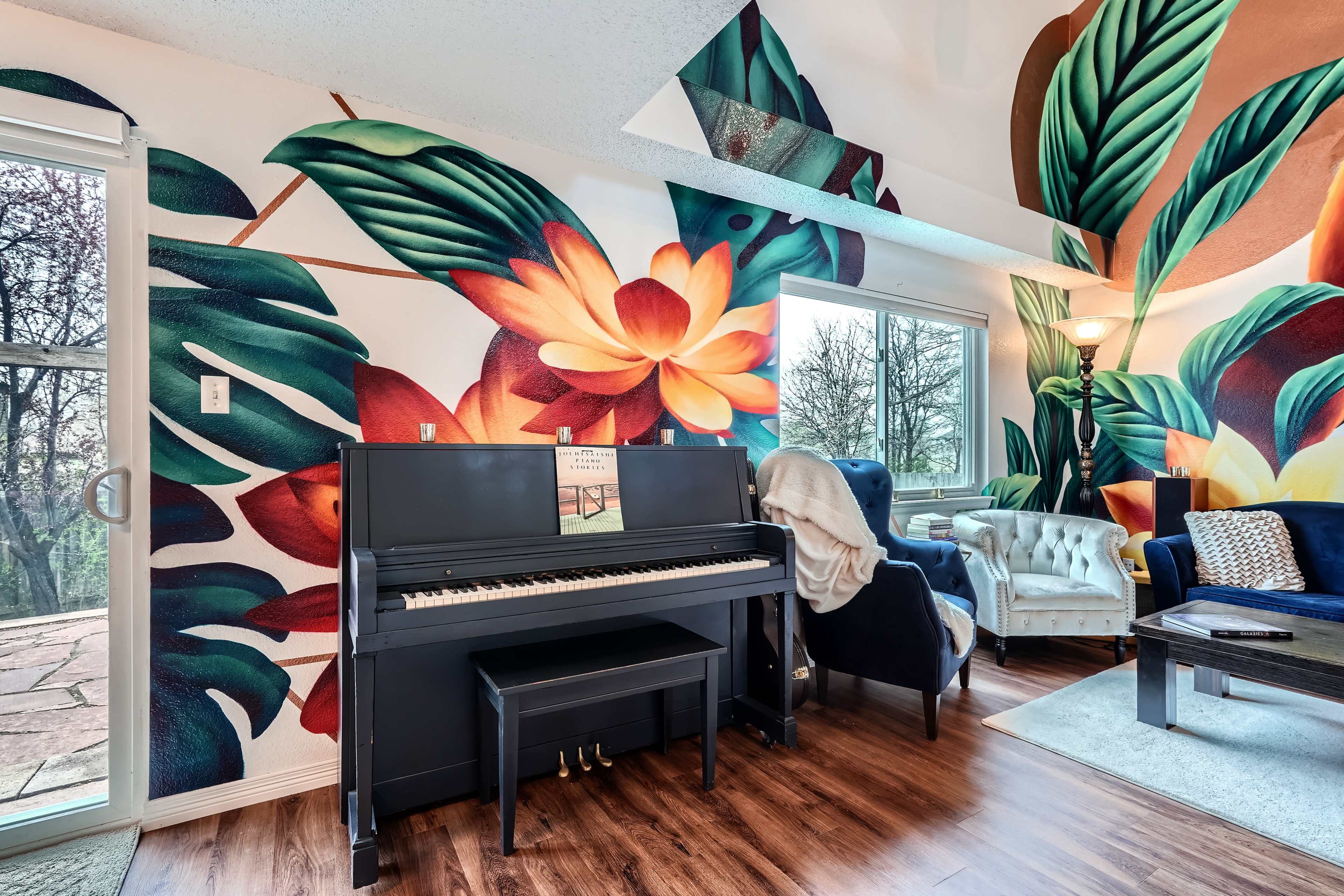 A black piano is positioned in front of a colorful mural featuring large leaves and flowers, with a window providing natural light to the room.