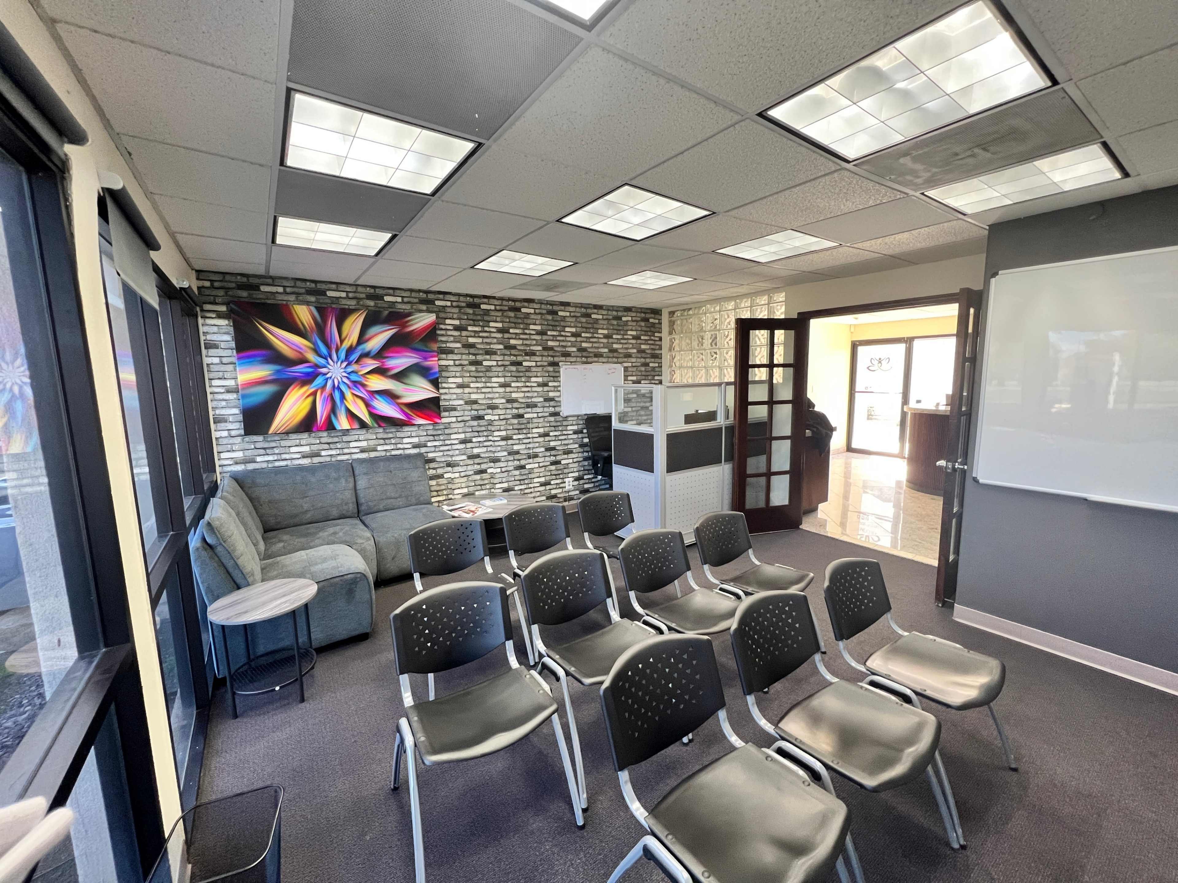 A modern office meeting room with a seating area featuring several chairs facing a presentation board, and a colorful artwork on the wall.
