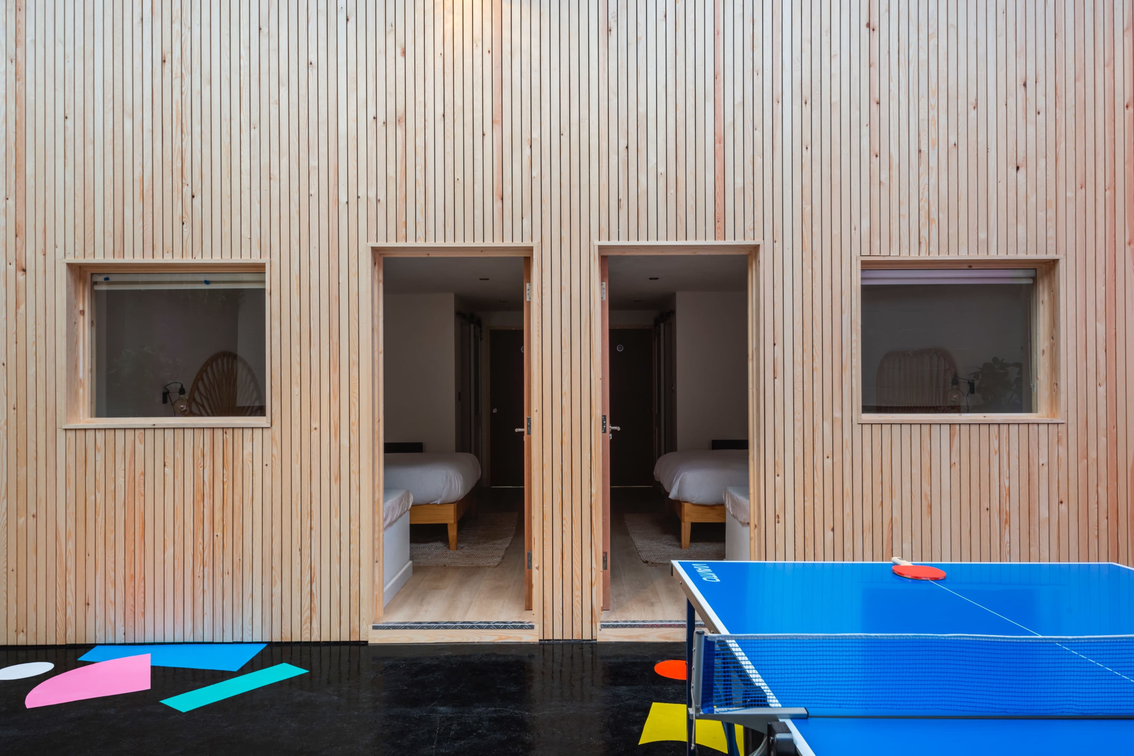 The image shows a wooden exterior of a building with two open doorways leading to separate rooms, and a blue ping pong table in the foreground with colorful shapes scattered on the floor.