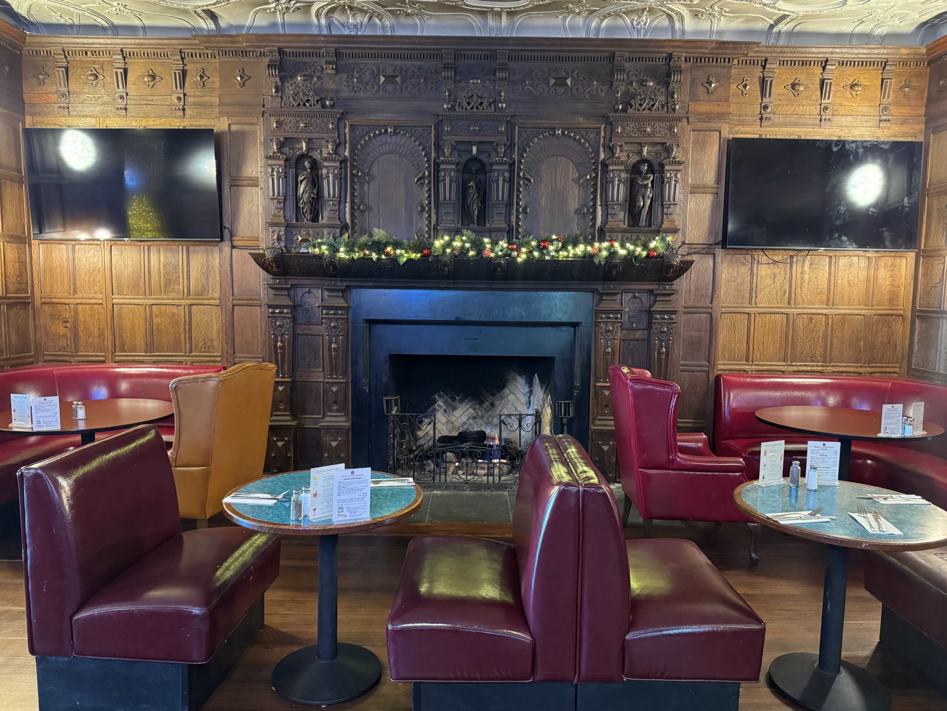 The image shows a cozy bar area with red and orange seating, a large wooden fireplace with decorative carvings, and two televisions mounted on the walls.