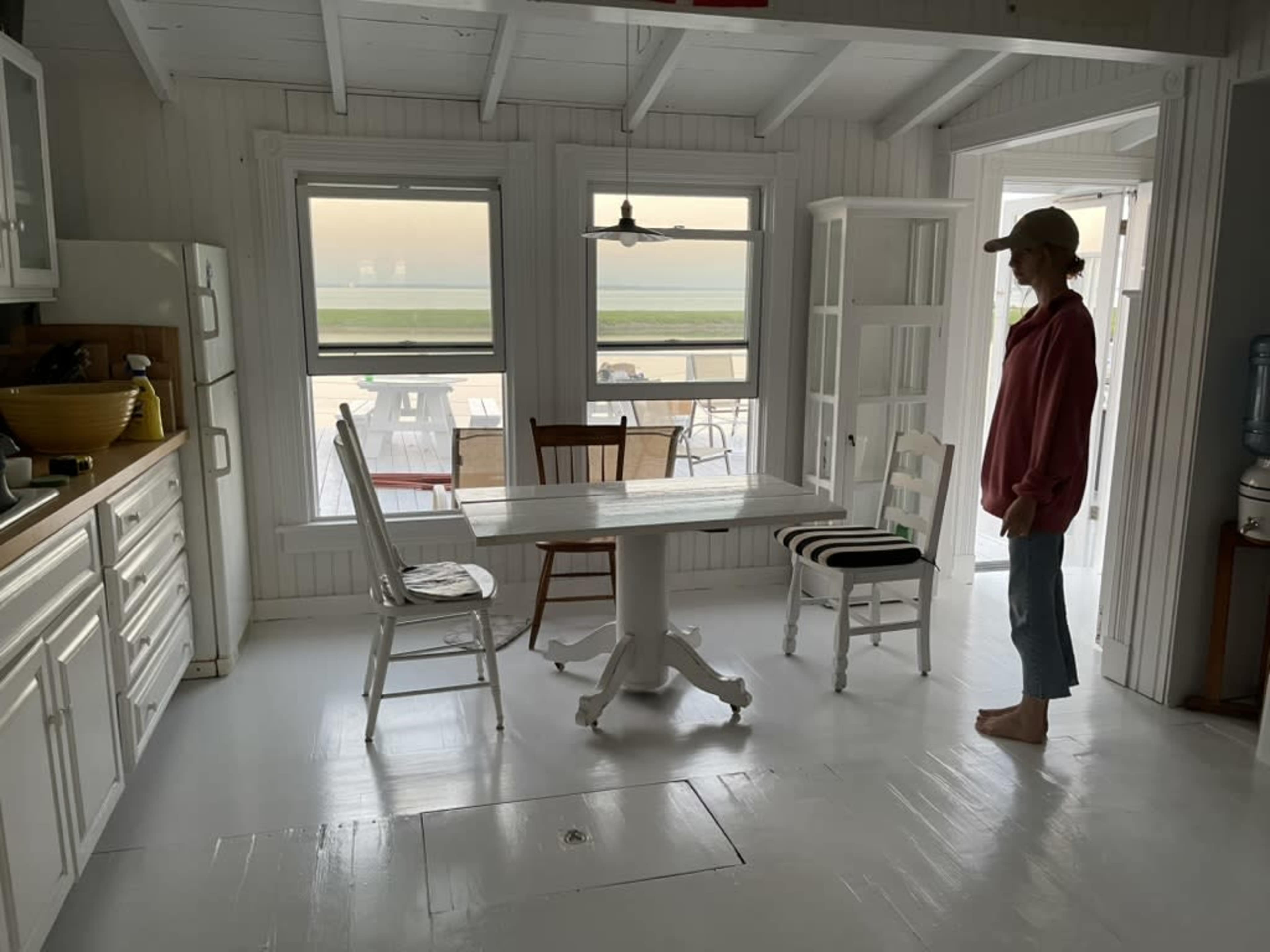 A person stands near a white dining table in a bright kitchen with large windows overlooking a patio area.