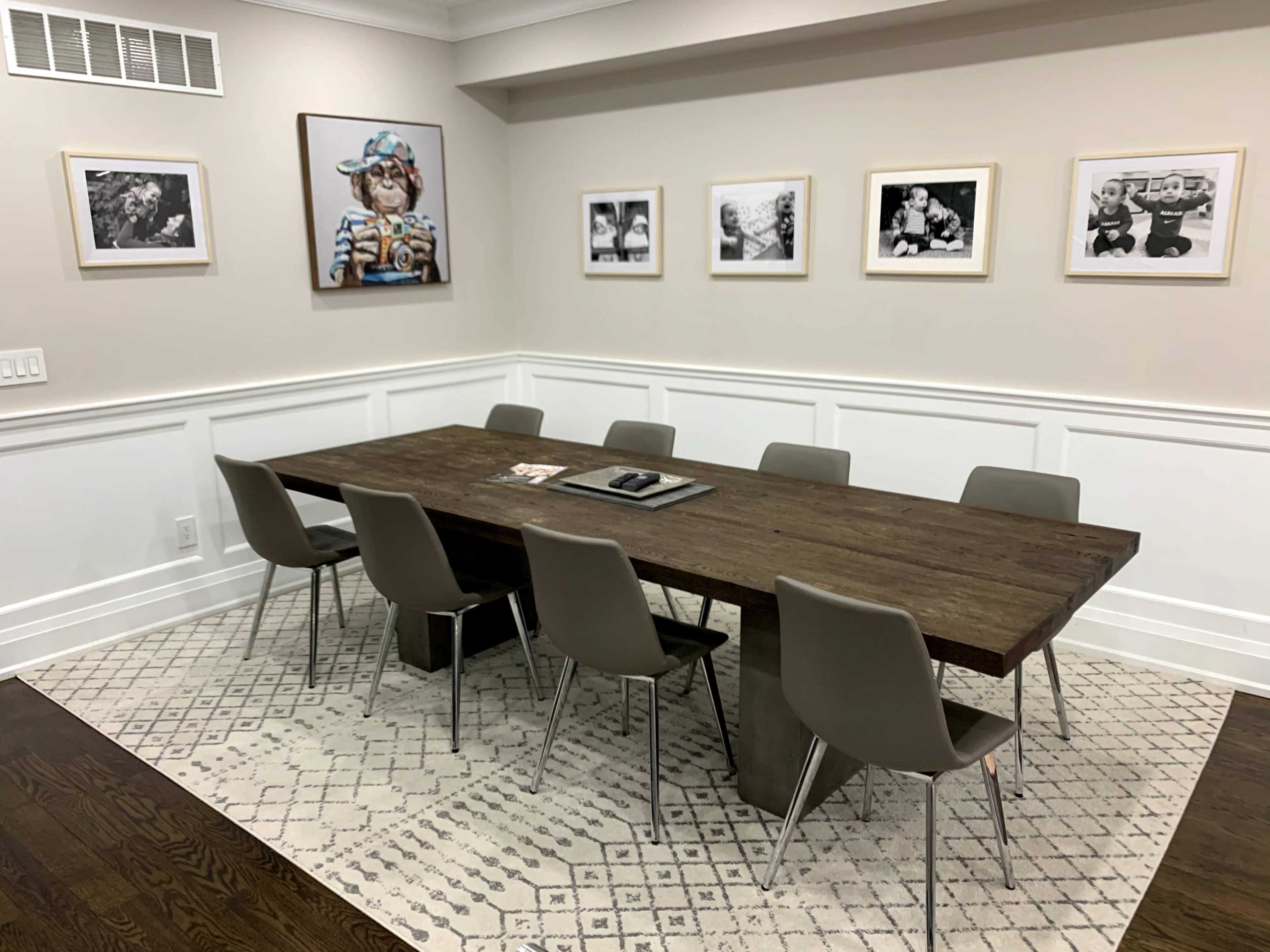 A rectangular wooden dining table is situated in a light-colored room with eight gray chairs and framed black-and-white photographs on the walls.