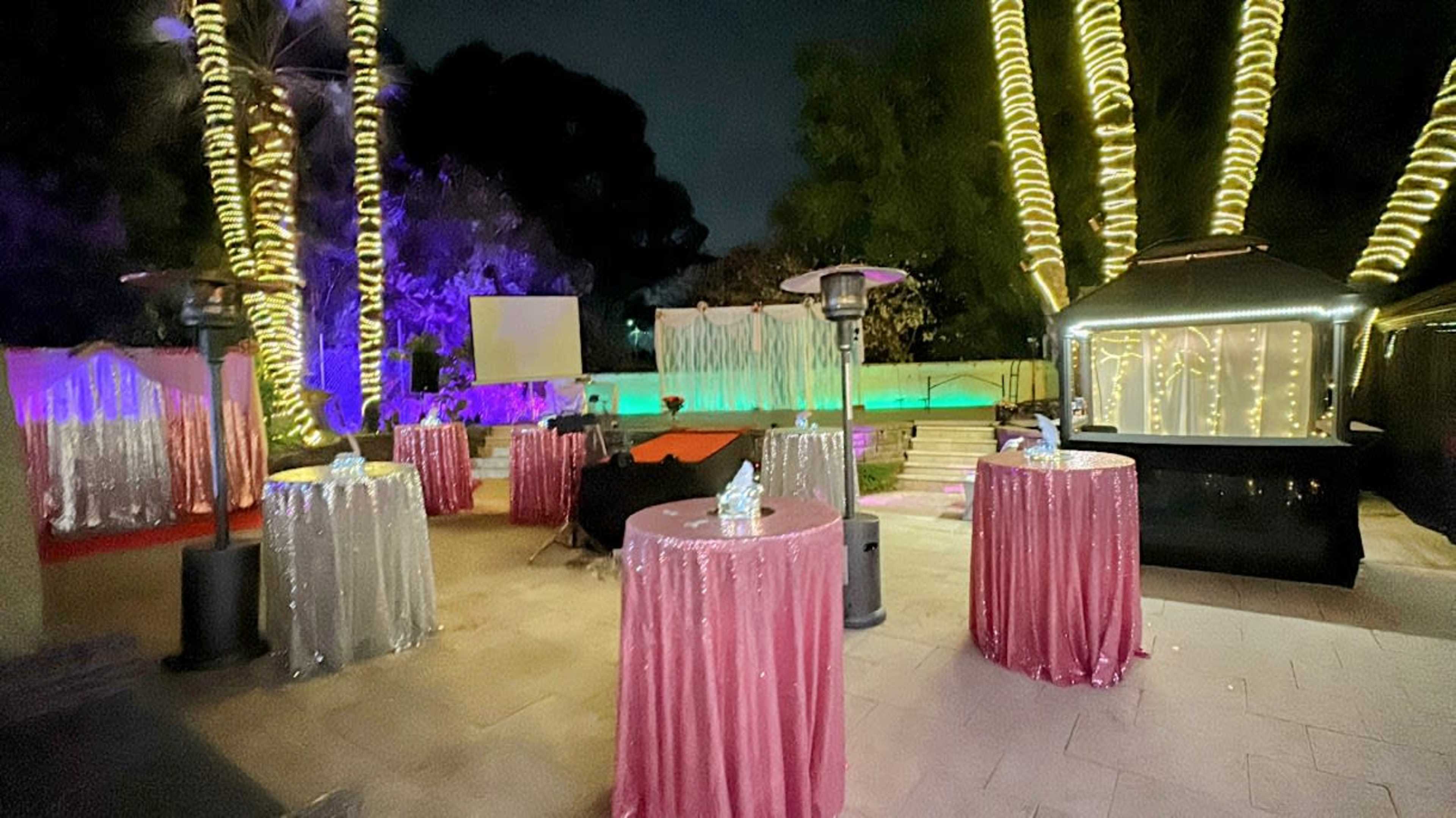 The scene depicts an outdoor event space at night, featuring tables with sparkling tablecloths, illuminated palm trees, and a backdrop of colorful lighting.