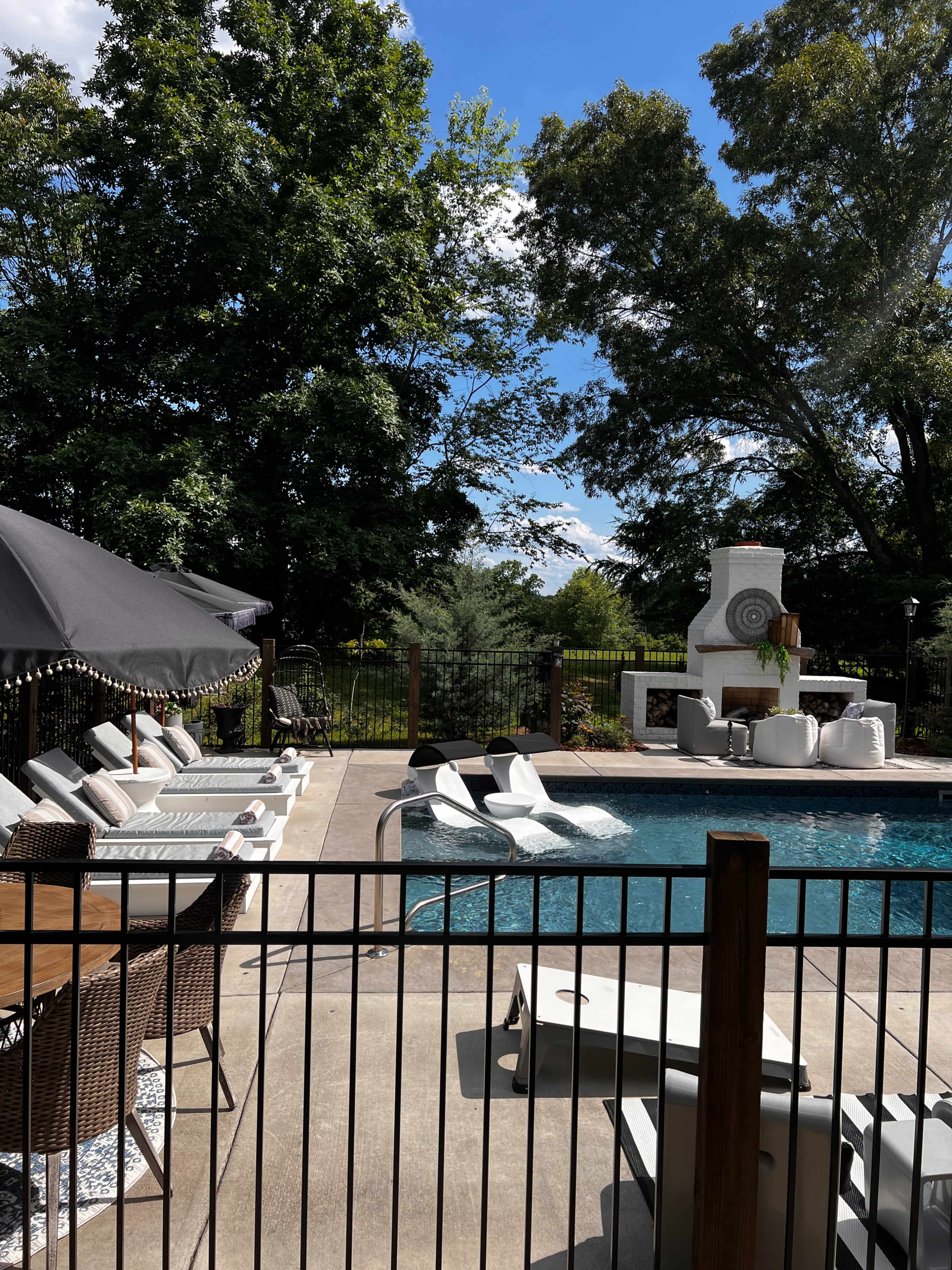 The image shows a pool area surrounded by a fence, featuring lounge chairs, an outdoor dining table, and a barbecue grill under a shaded umbrella.