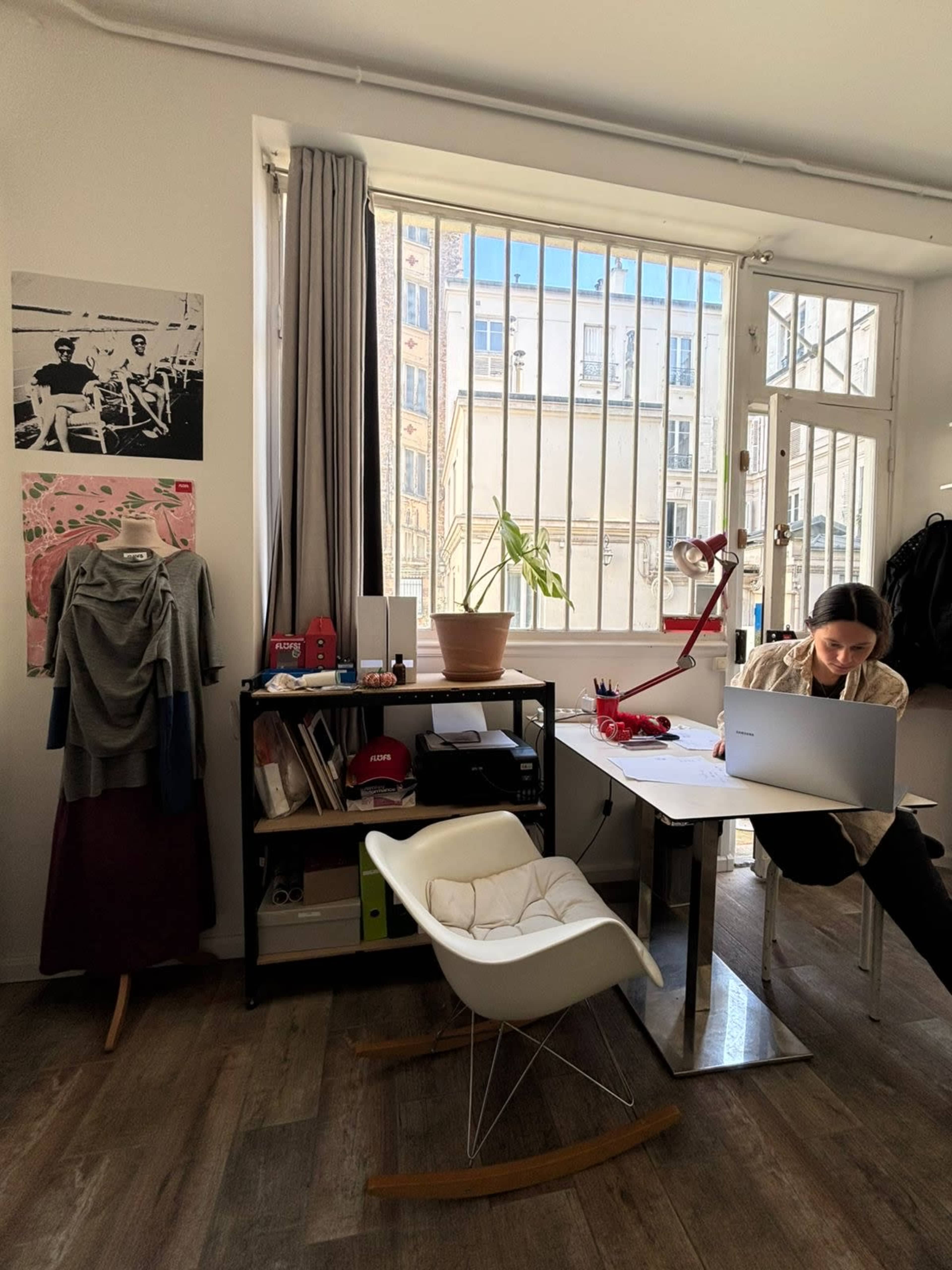 A person is seated at a desk working on a laptop in a bright room with large windows and various items on display.
