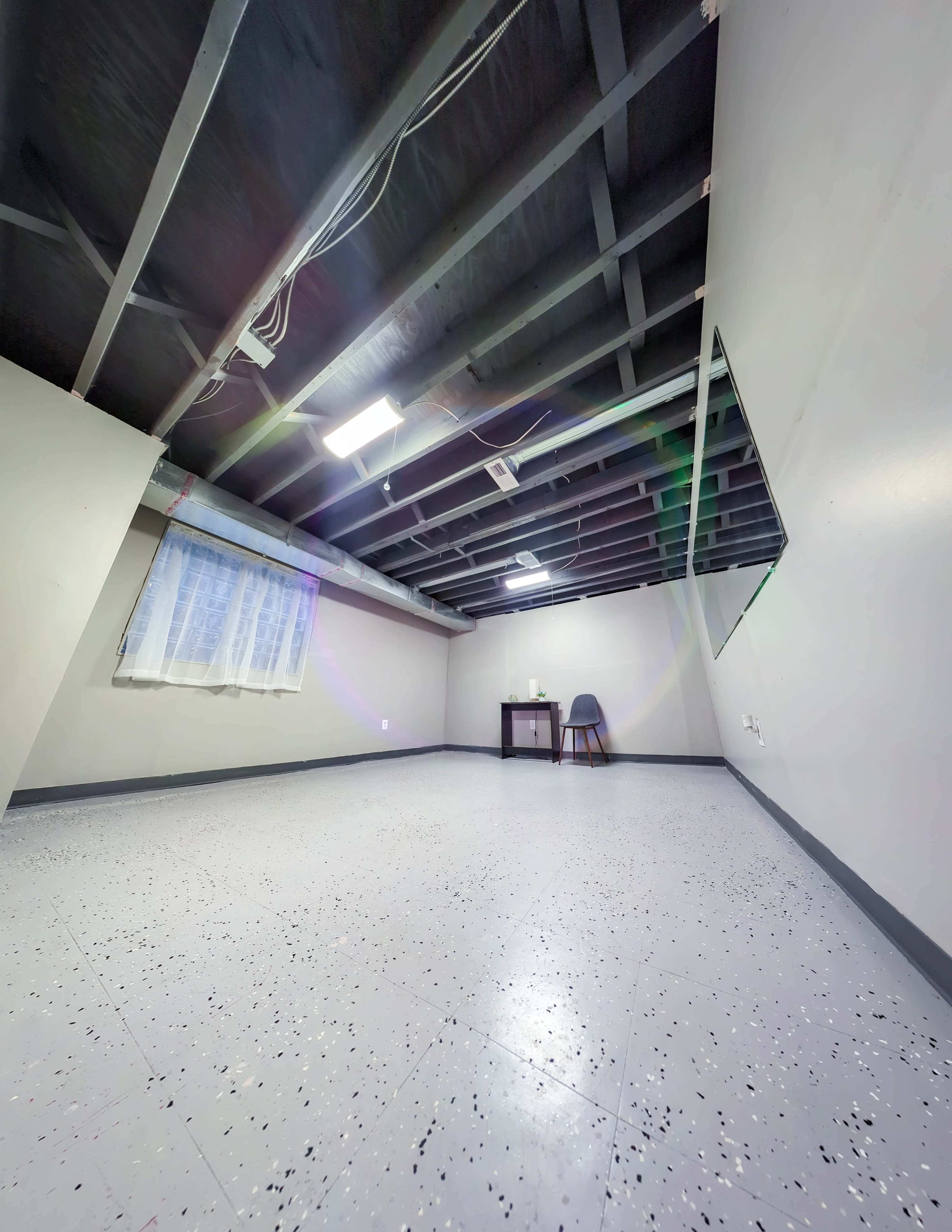 The image shows a sparsely furnished room with a bare concrete floor, exposed ceiling, and a small table with a chair in one corner.
