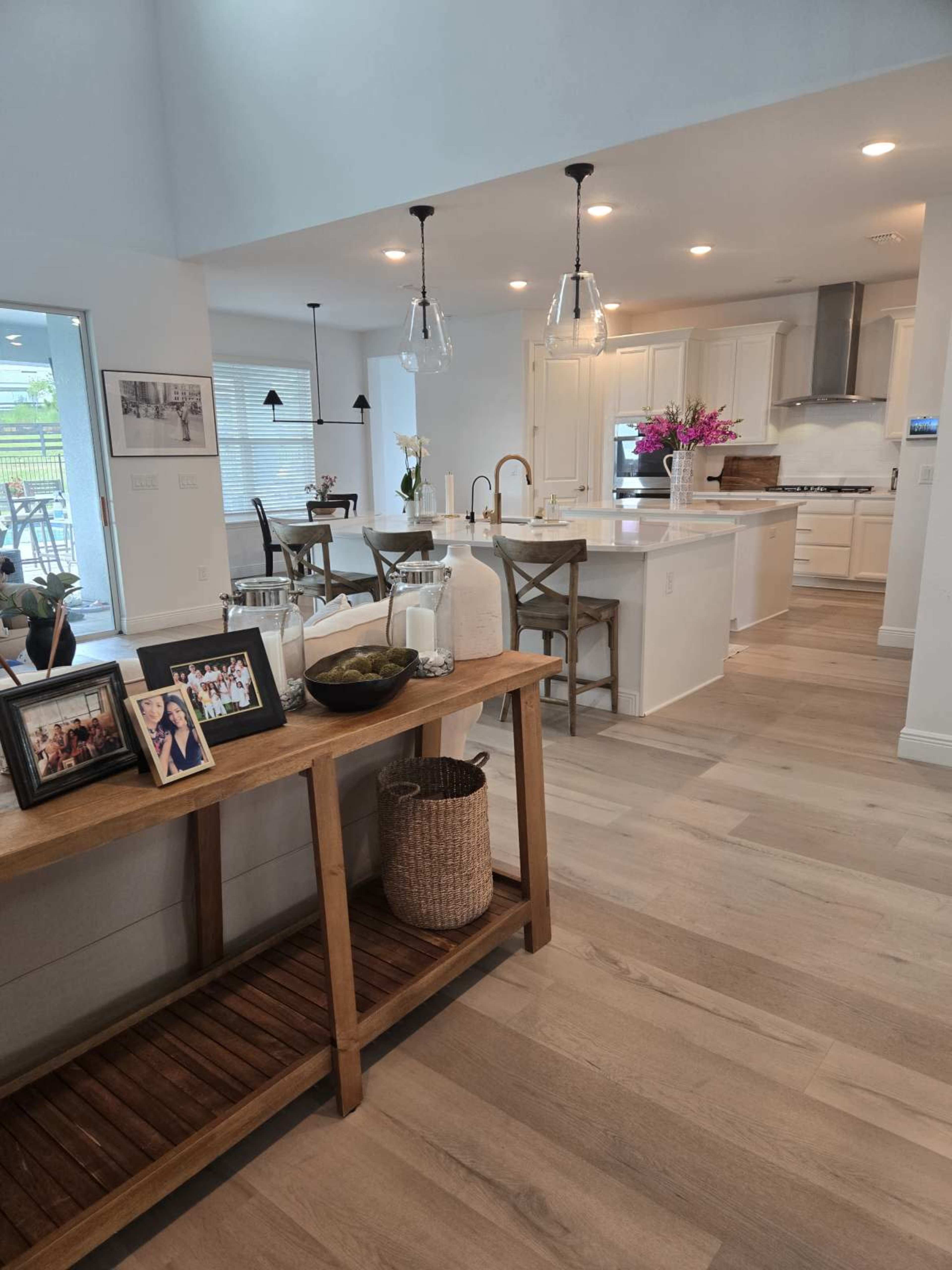 The image shows a modern open-concept kitchen and dining area with light wood floors, white cabinetry, and a wooden console table displaying decorative items and framed photos.