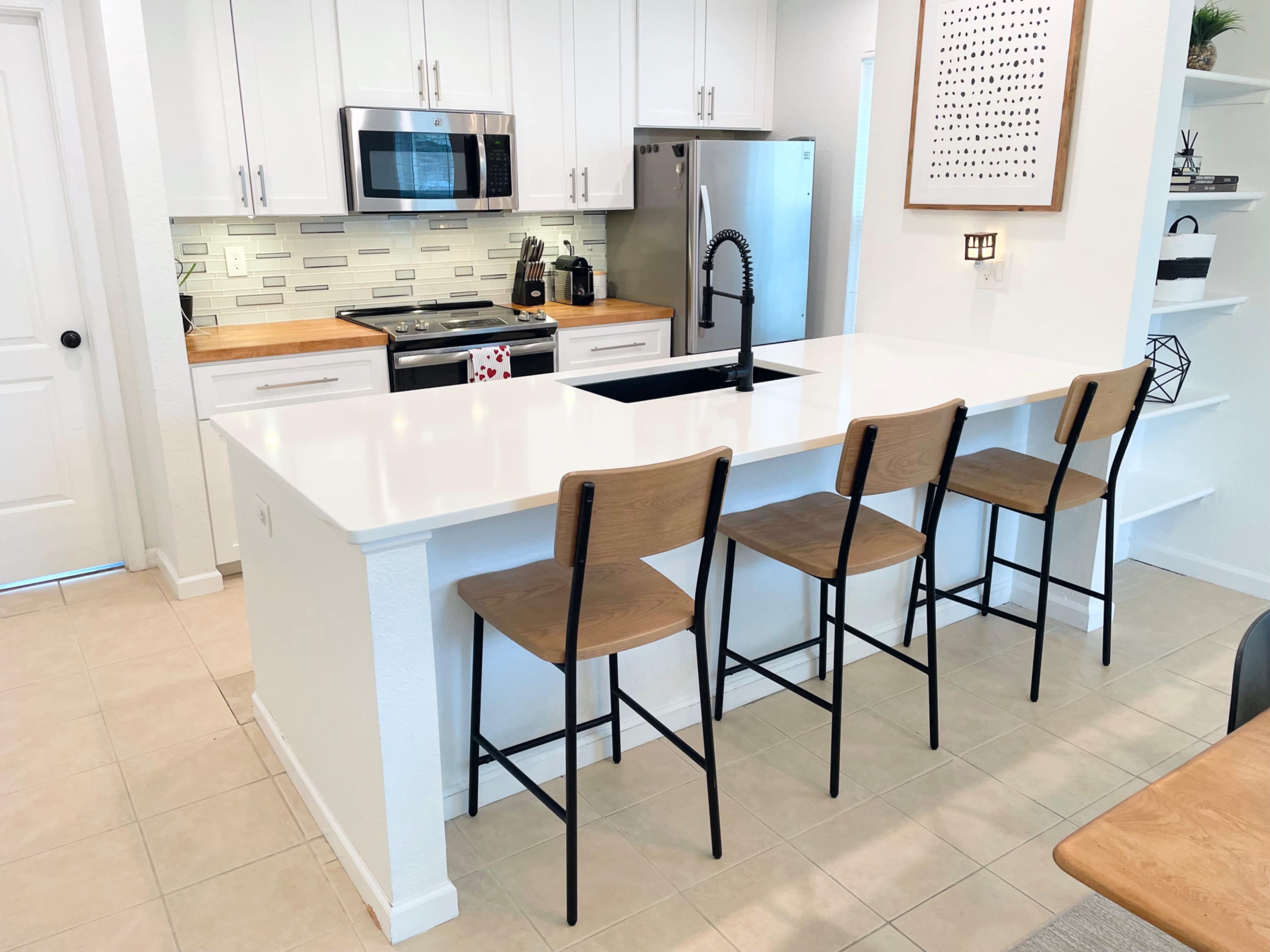 A modern kitchen features white cabinetry, stainless steel appliances, a large island with a sink, and four wooden bar stools.