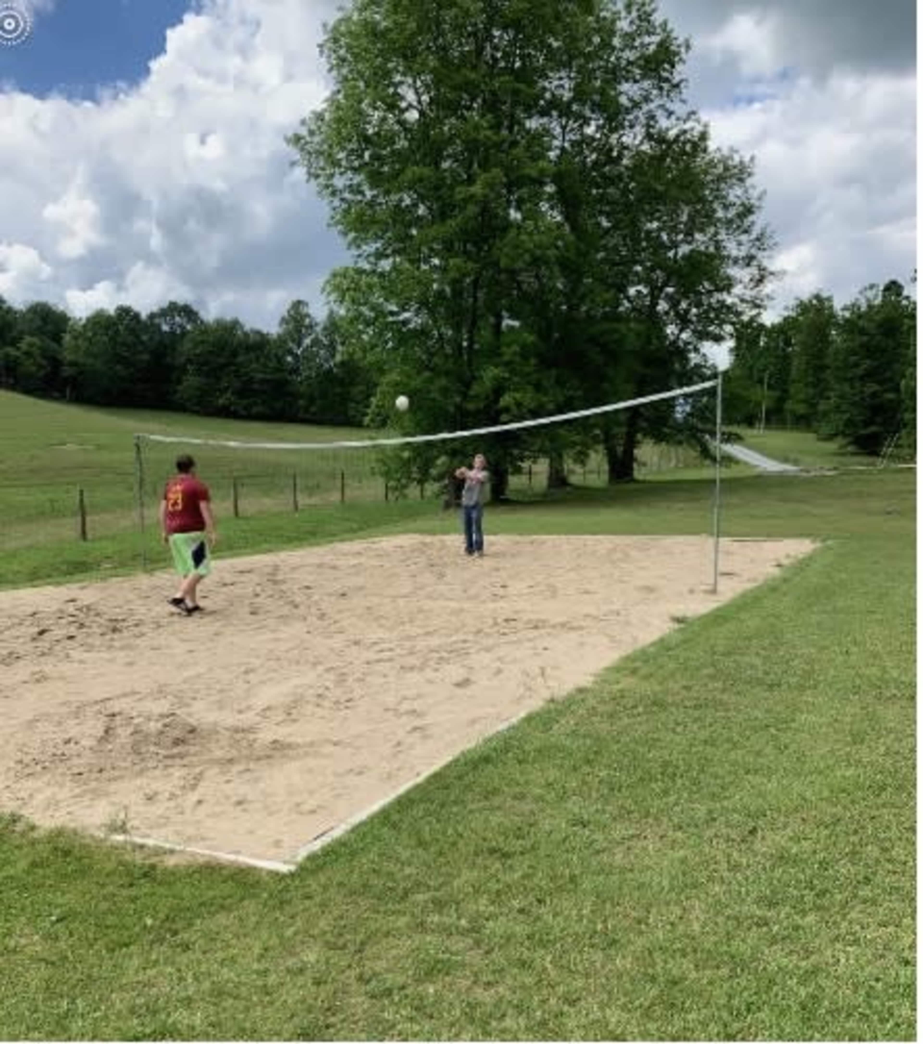 Two people are playing volleyball on a sand court surrounded by grass and trees under a partly cloudy sky.