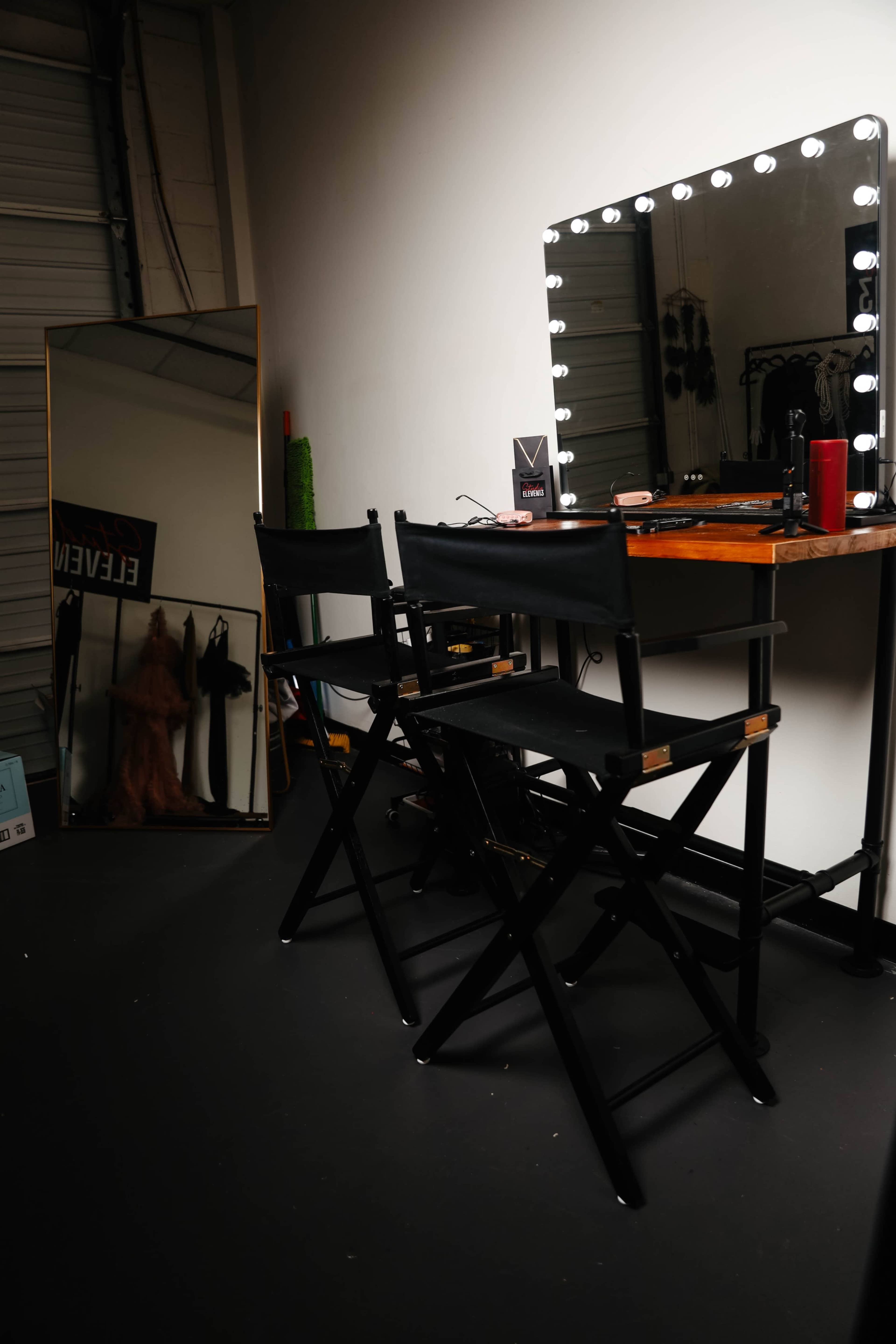 The image shows a vanity setup with two black director's chairs, a lighted mirror, and a wooden table in a dimly lit room with a large mirror reflecting the scene.