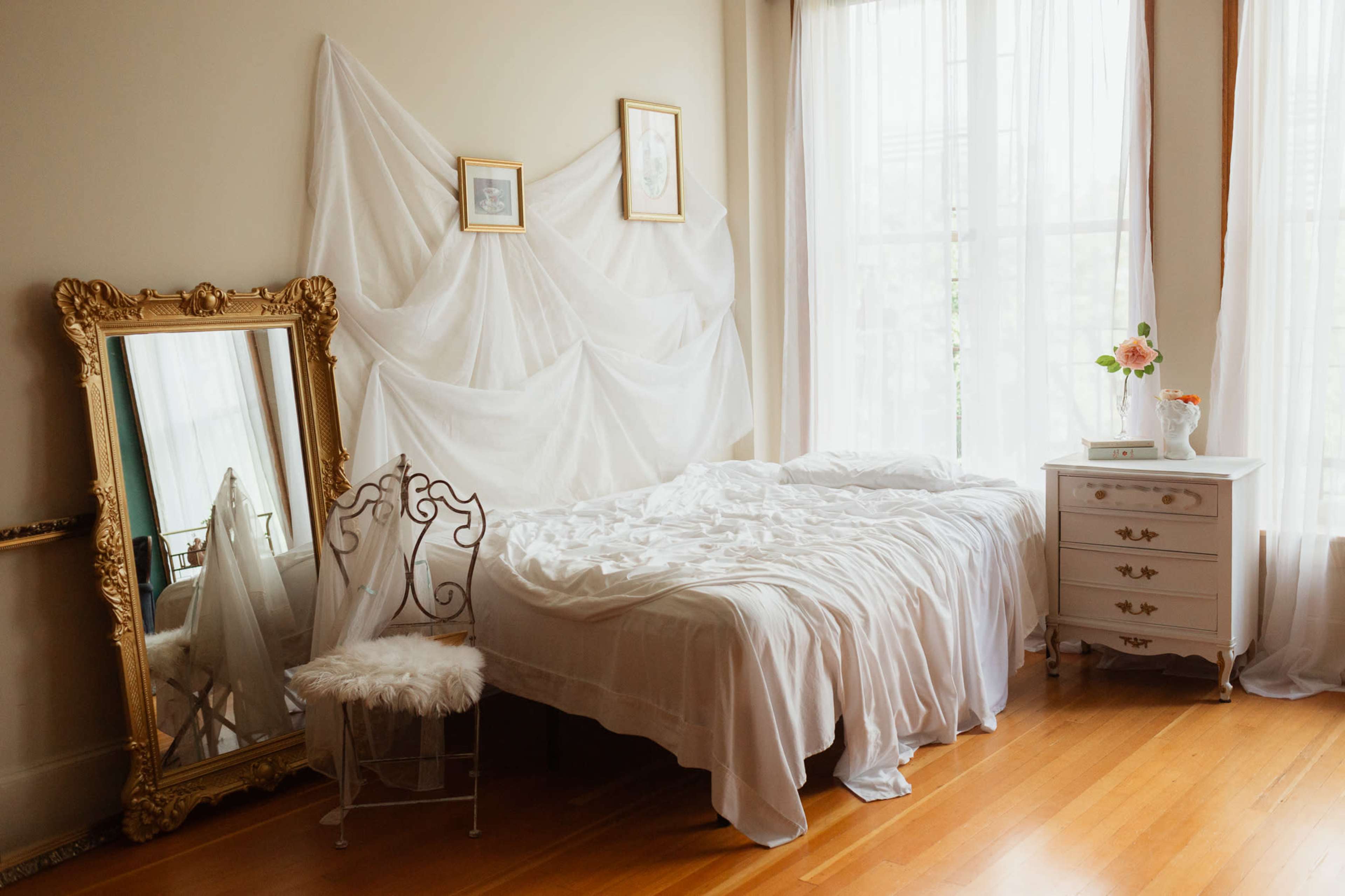 A neatly made bed with white linens is positioned next to a white nightstand and a tall mirror in a bright room adorned with sheer drapes.