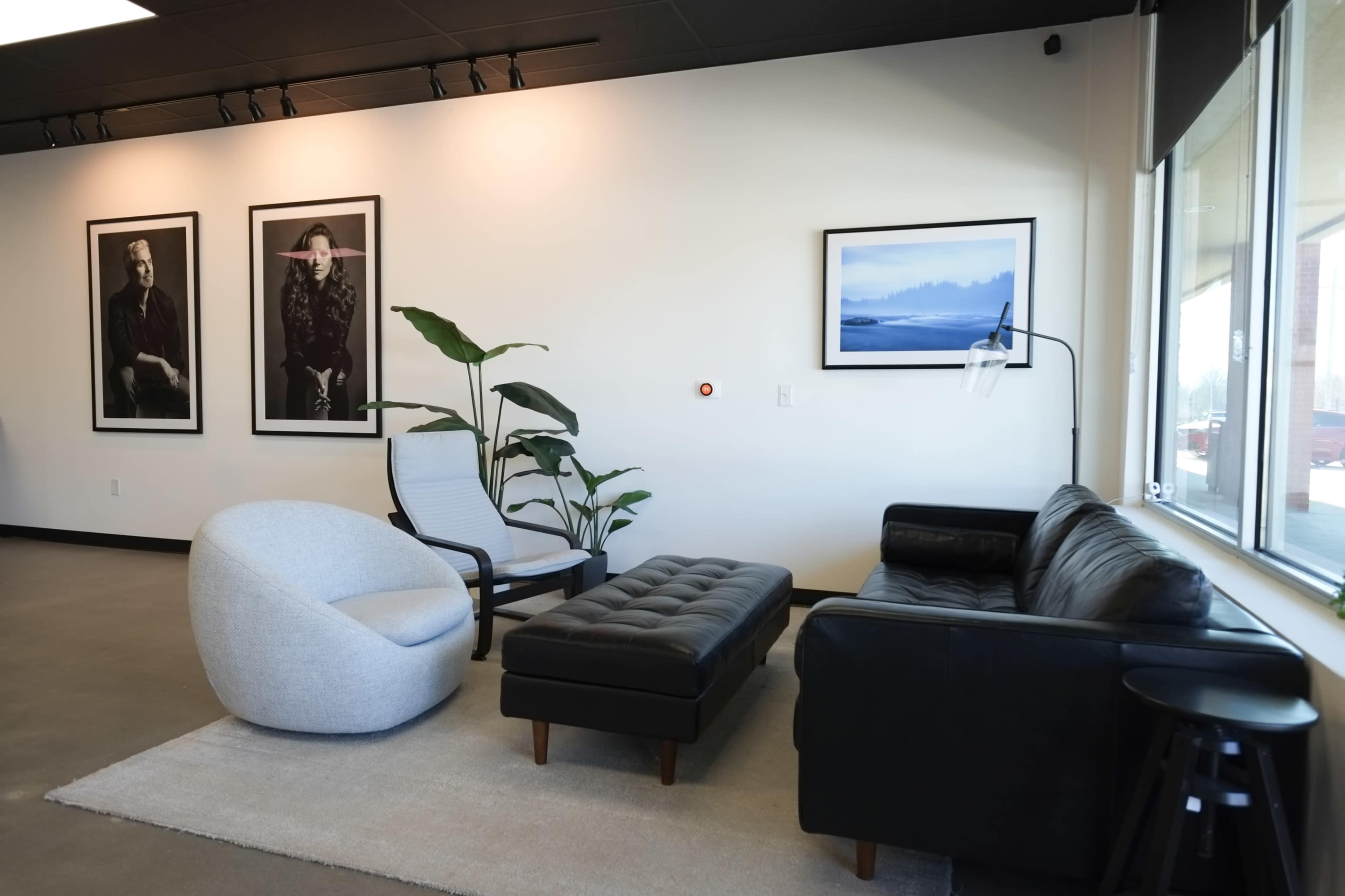 The image shows a modern lounge area with a black leather sofa, a fabric armchair, and a tufted ottoman, framed by large photographs on the wall and a potted plant nearby.