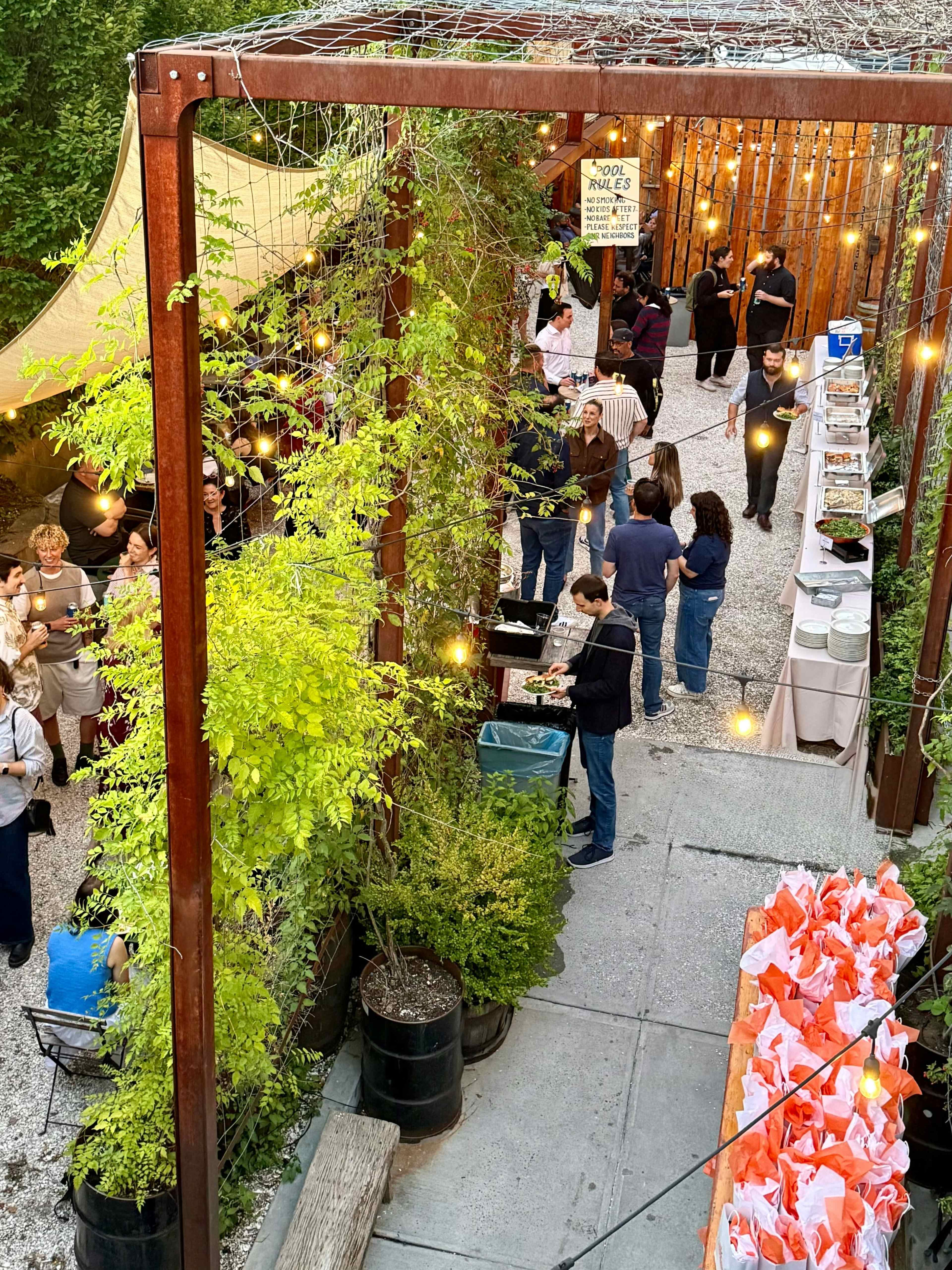 An outdoor event space features string lights and decorated tables, with people mingling and enjoying food along a gravel path surrounded by greenery.