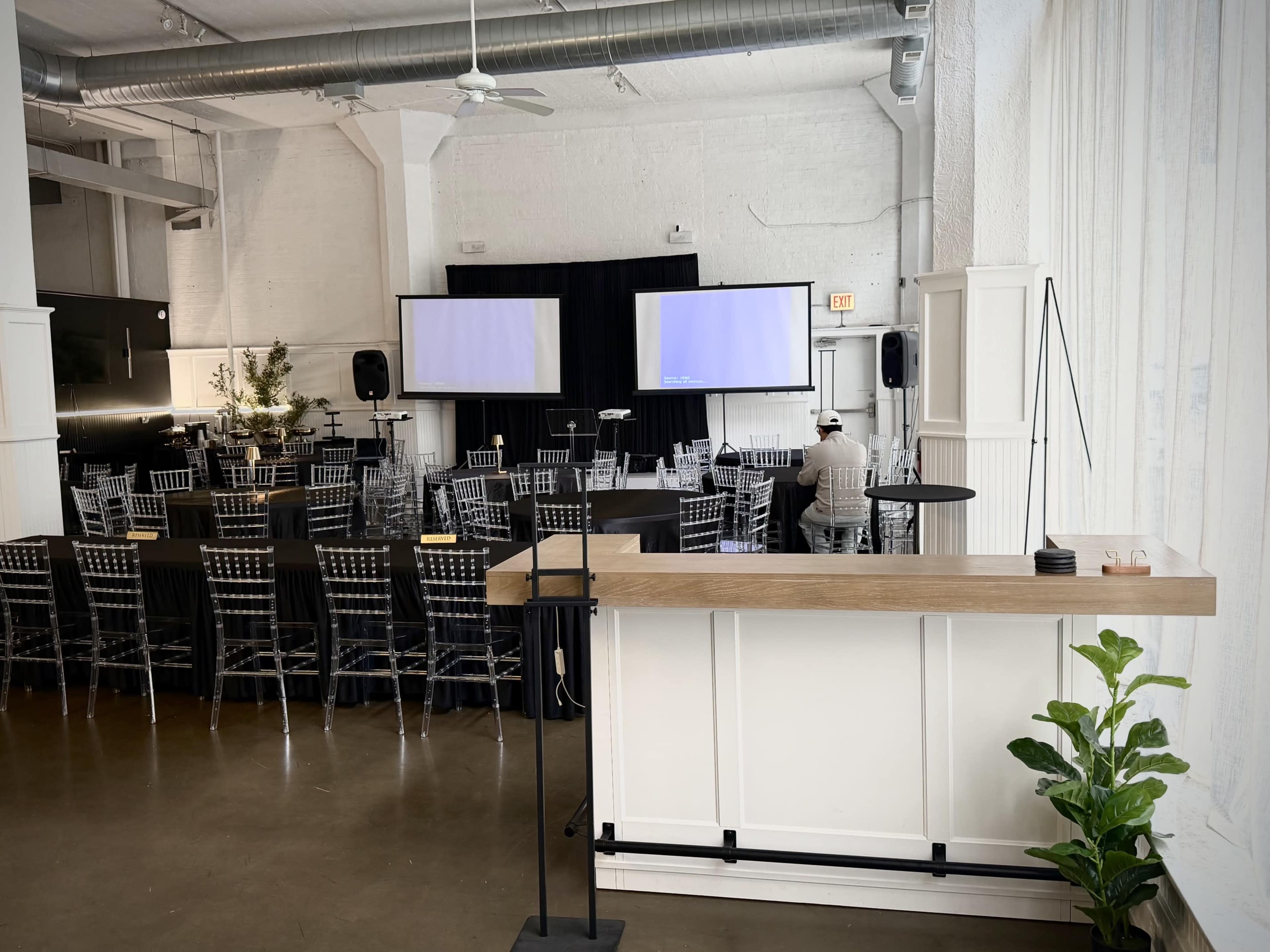 A modern event space features rows of black chairs and two large screens at the front, with a bar area in the foreground and a potted plant next to it.
