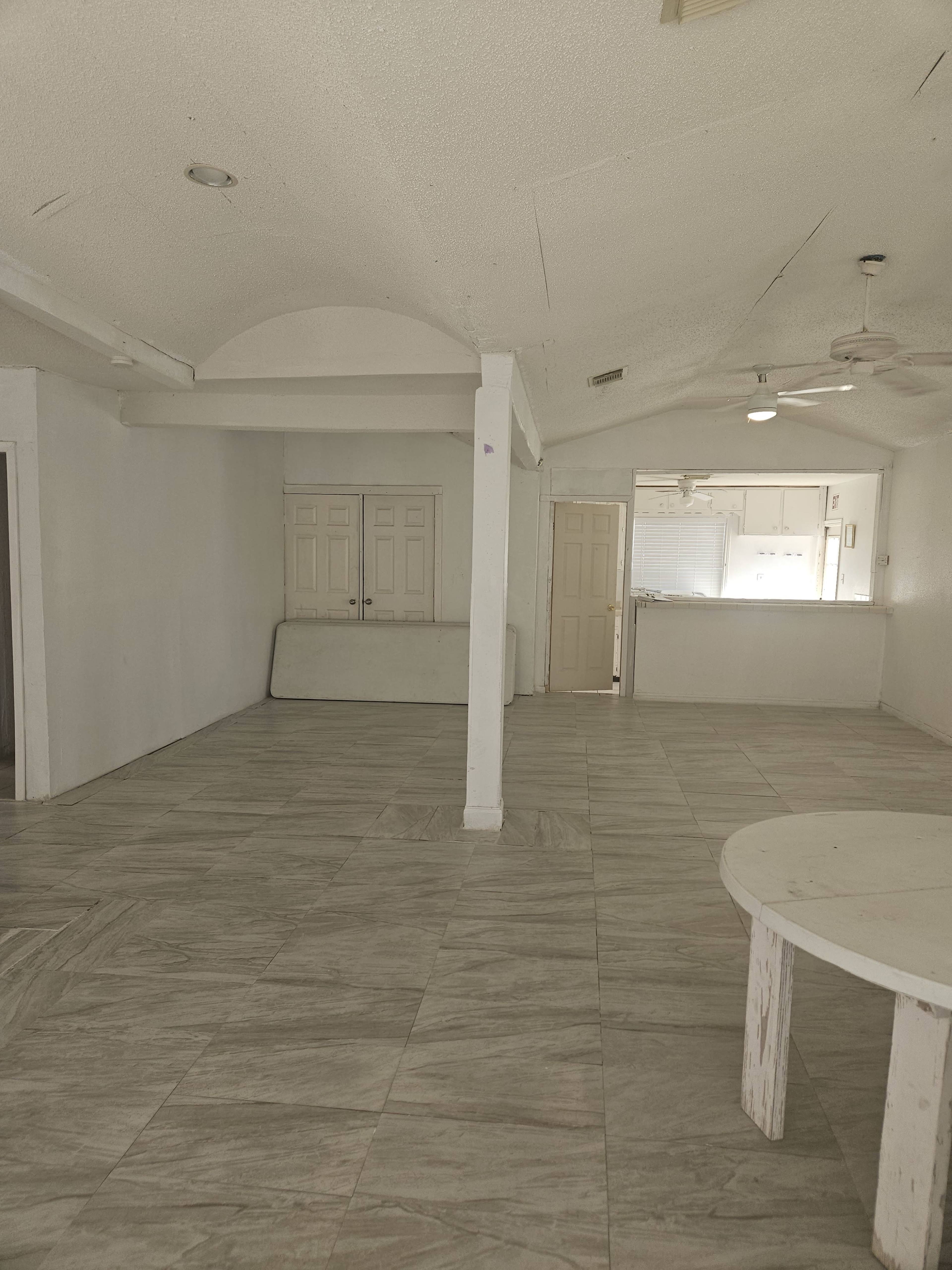 The image shows a spacious, empty room with tiled flooring, a central support column, and an adjoining area that appears to lead into a kitchen.