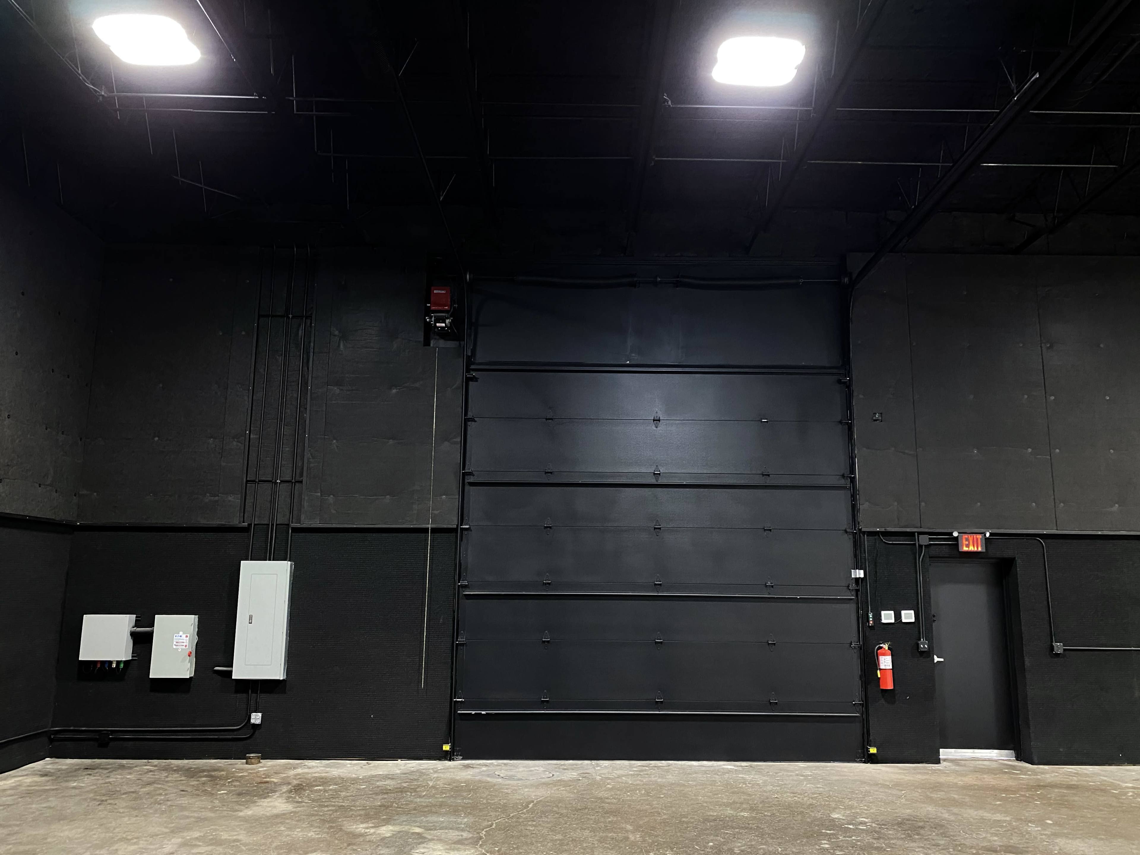 An empty industrial space with a large black roll-up door, electrical panels, and a fire extinguisher near a door in the corner.