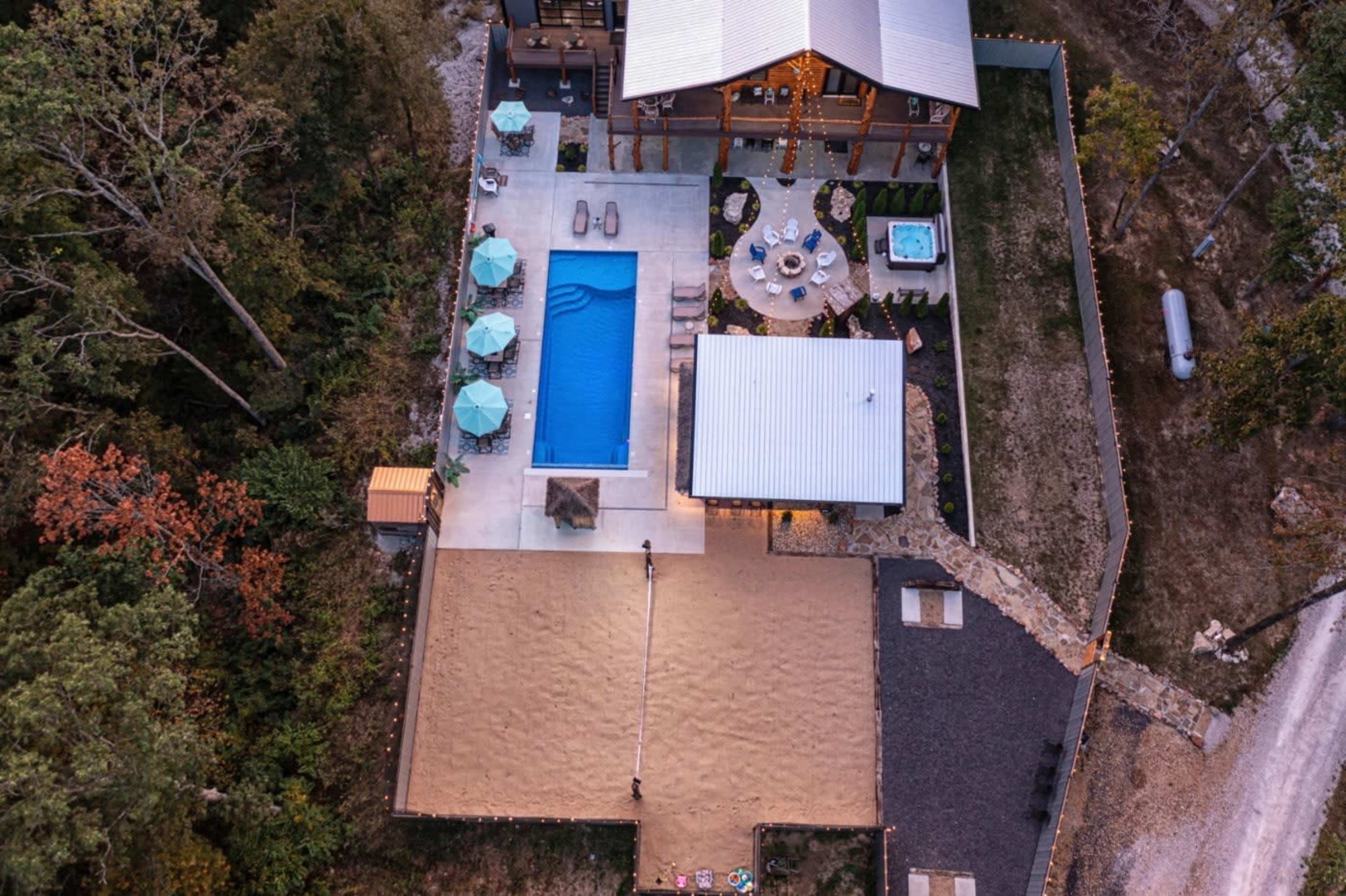 An aerial view of a property featuring a swimming pool, a hot tub, outdoor seating areas, and a sand volleyball court, surrounded by trees.