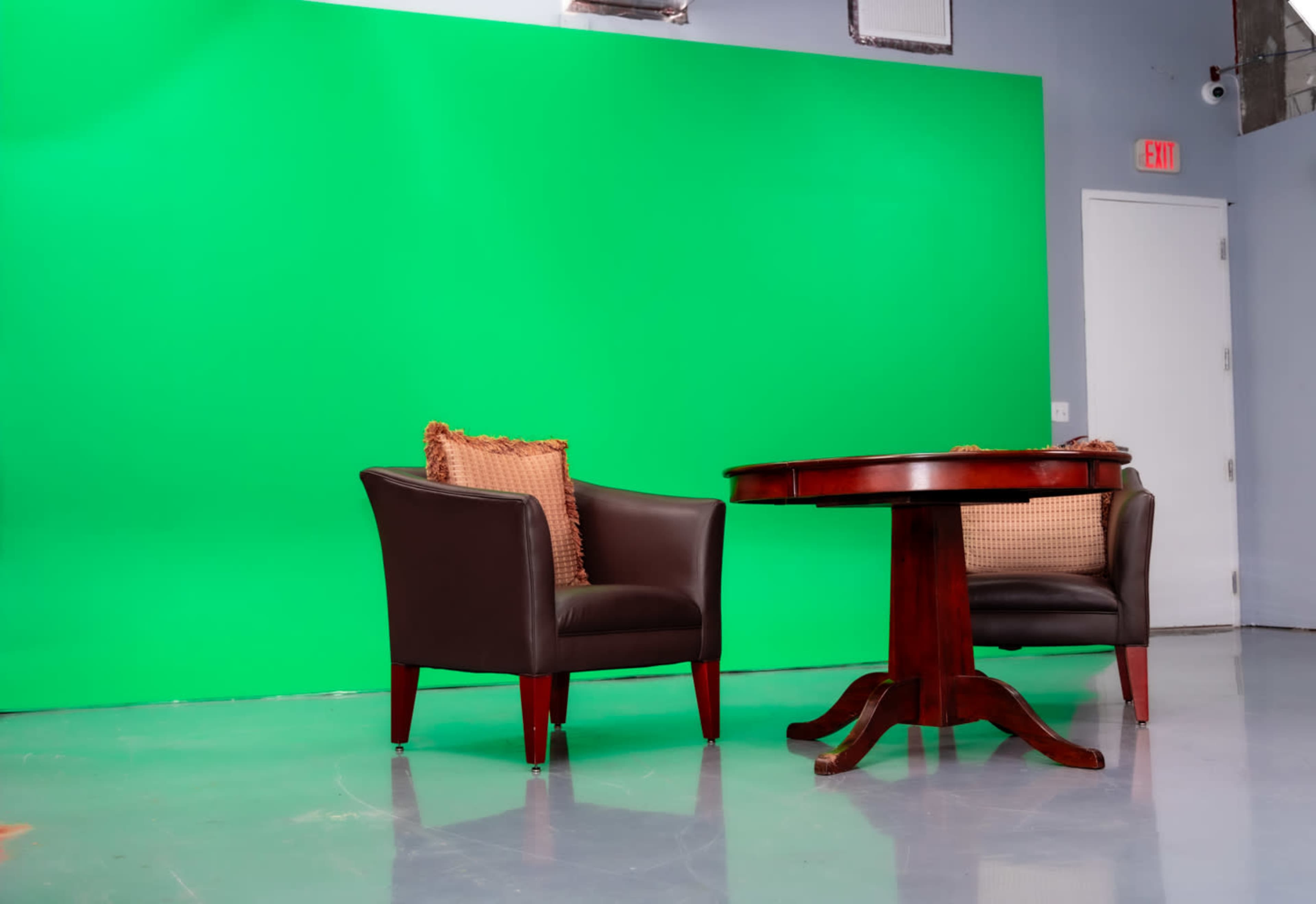 The image shows two dark leather chairs with decorative cushions positioned around a wooden table in front of a green screen.