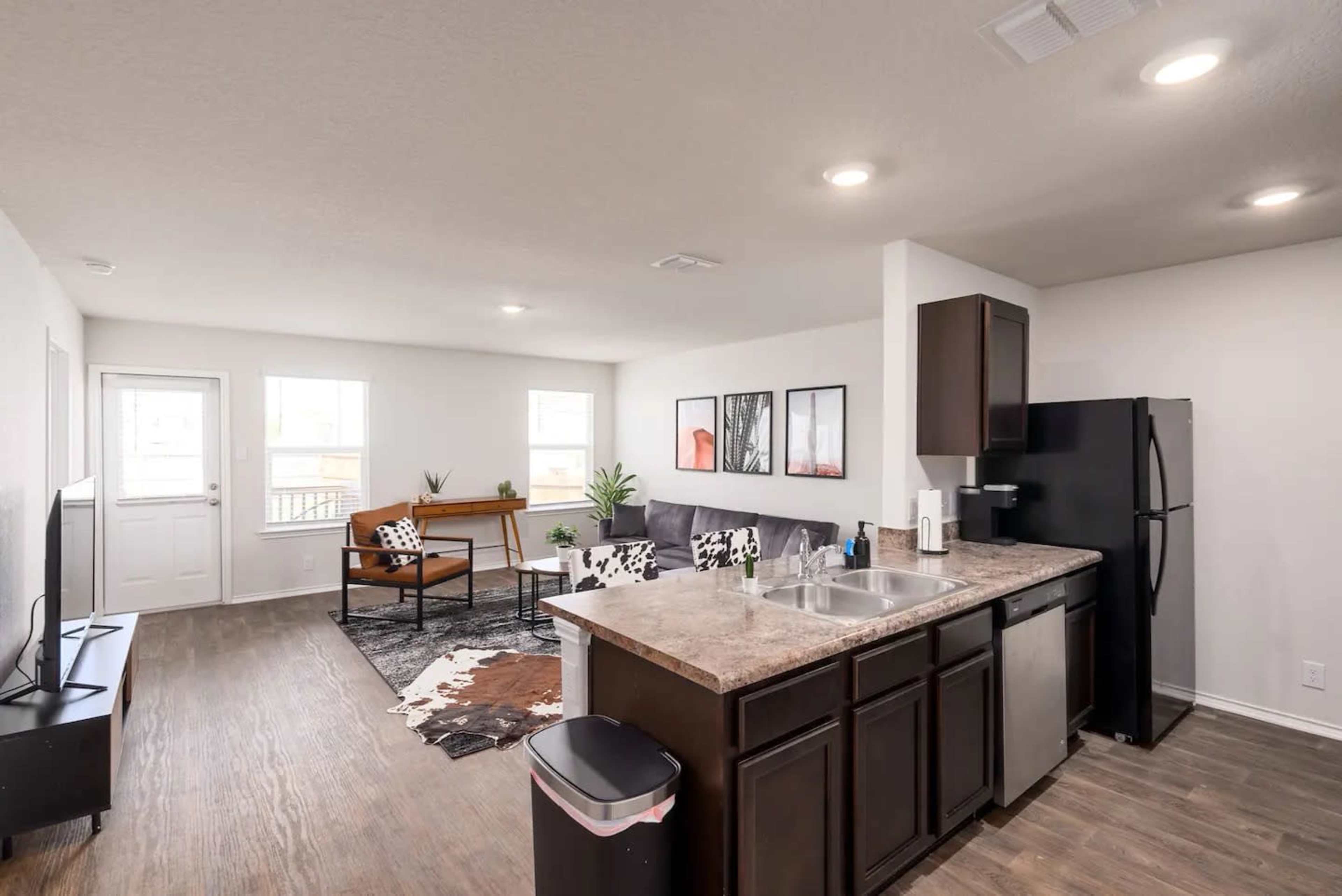 A modern living room and kitchen area with dark cabinetry, a black refrigerator, a sofa, and a seating arrangement, all illuminated by overhead lighting.