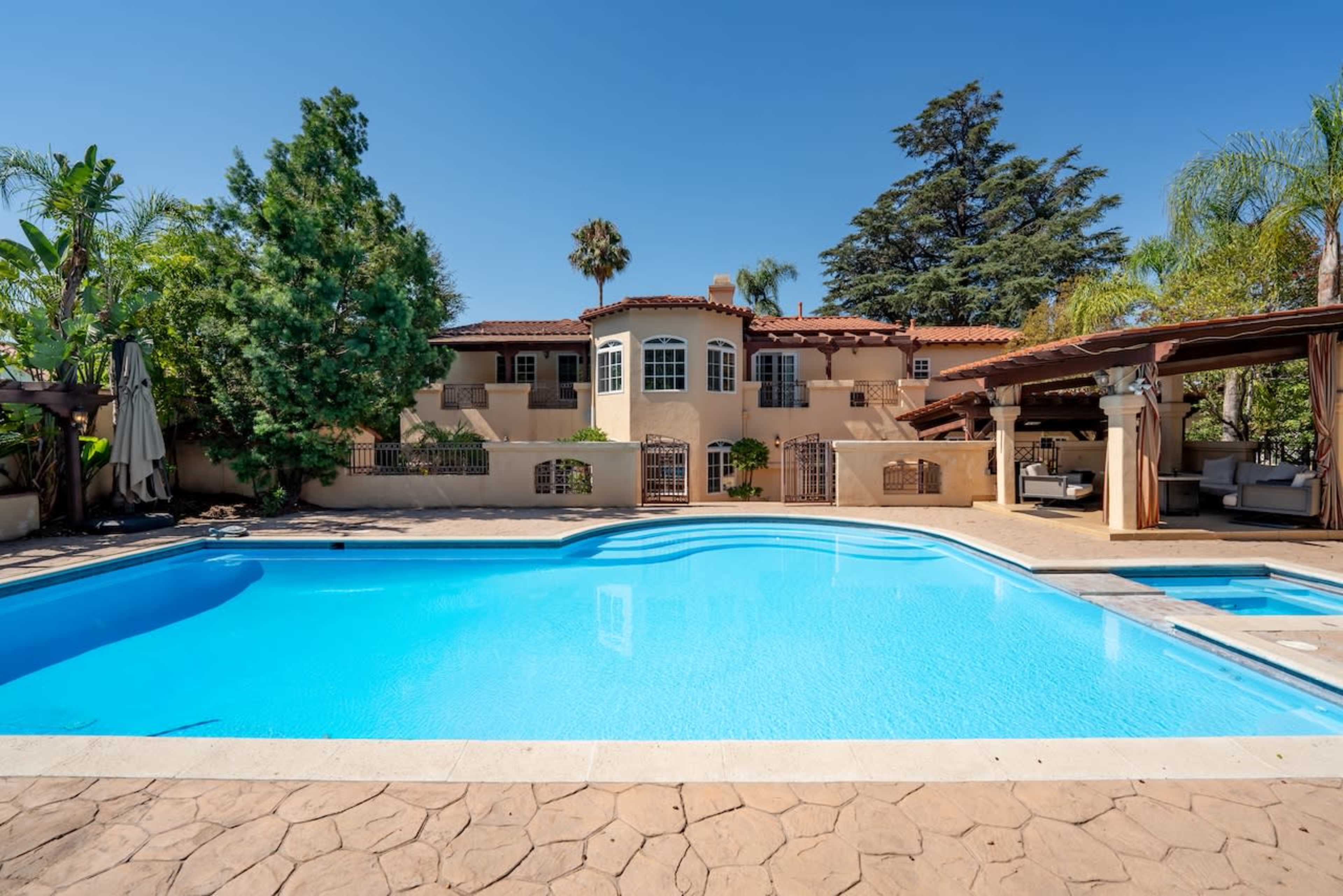 A large, clear swimming pool is surrounded by palm trees and a two-story house with multiple windows in the background.