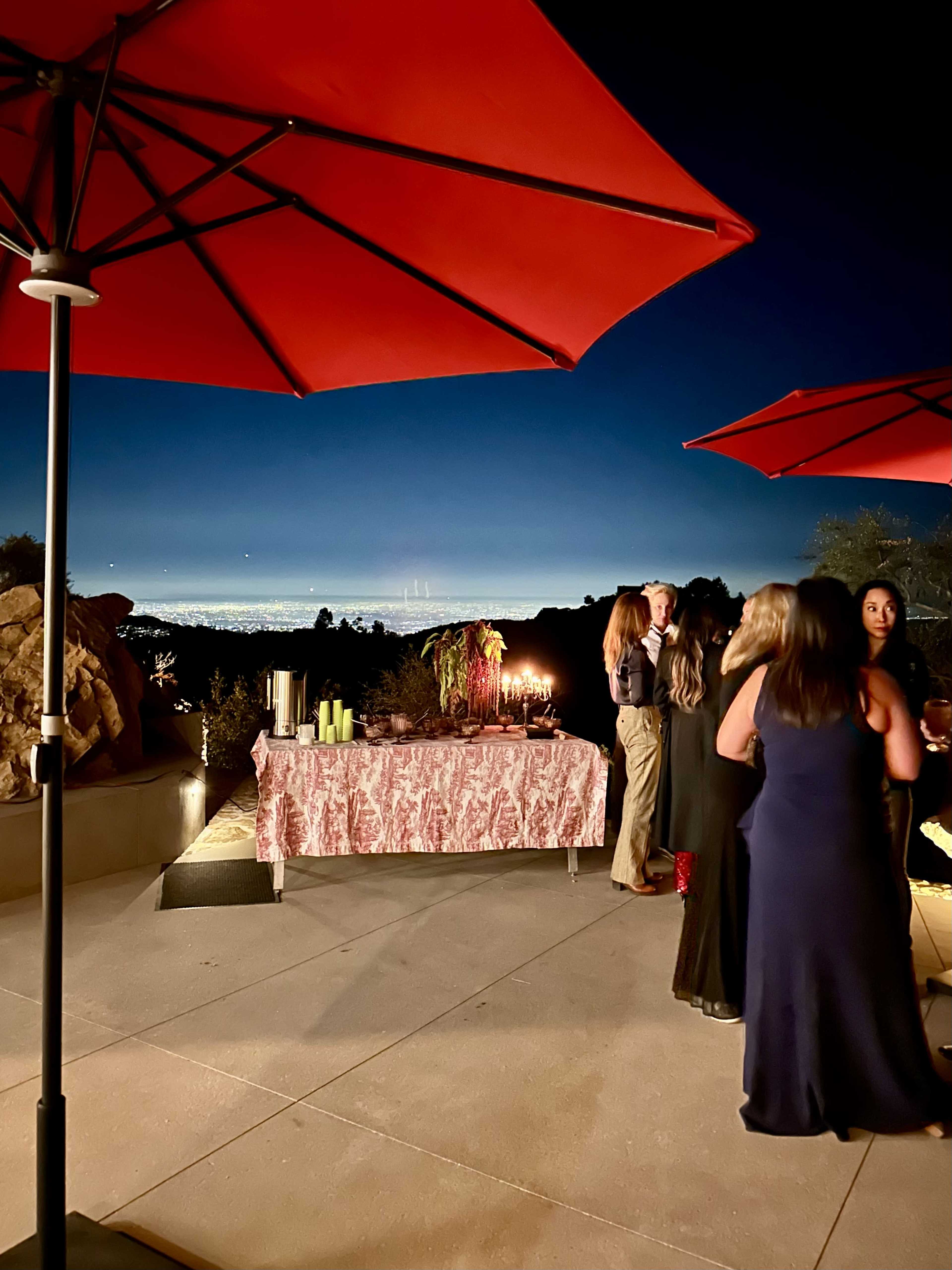 A group of people gathers near a buffet table under red umbrellas, overlooking a cityscape at dusk.