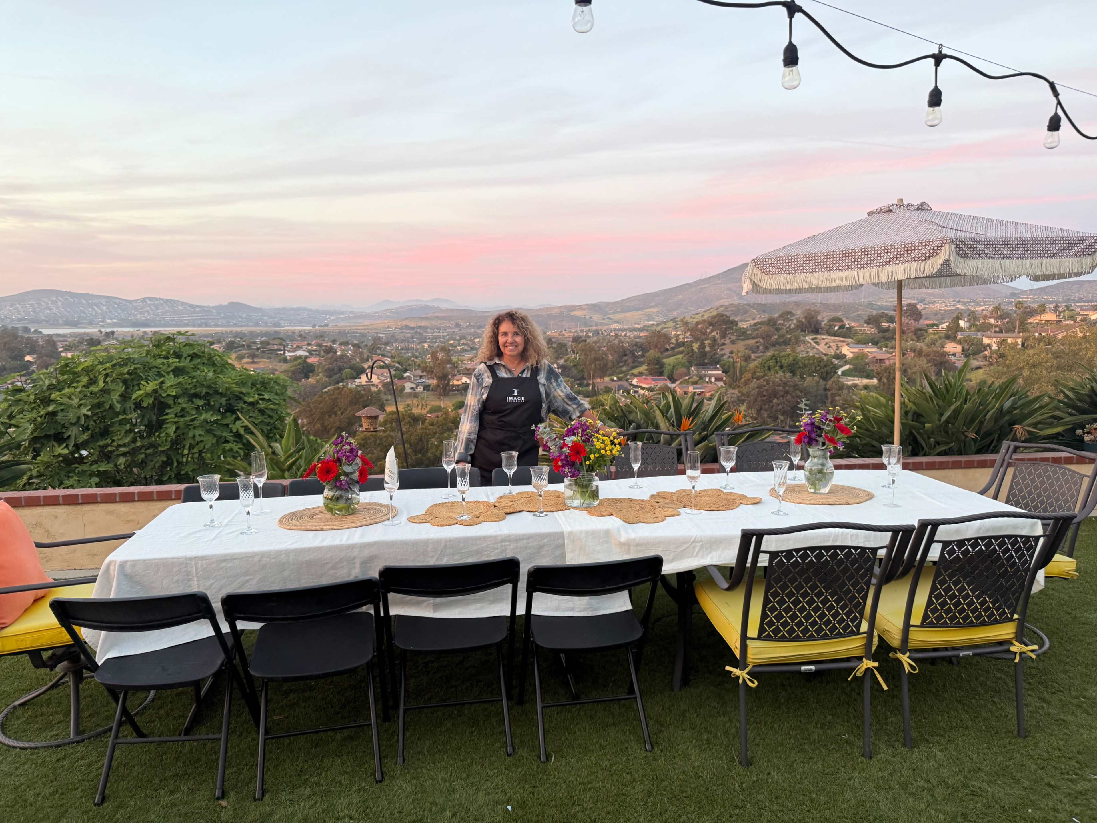 A woman stands beside a long dining table set outdoors with a view of a landscape under a pink sky.
