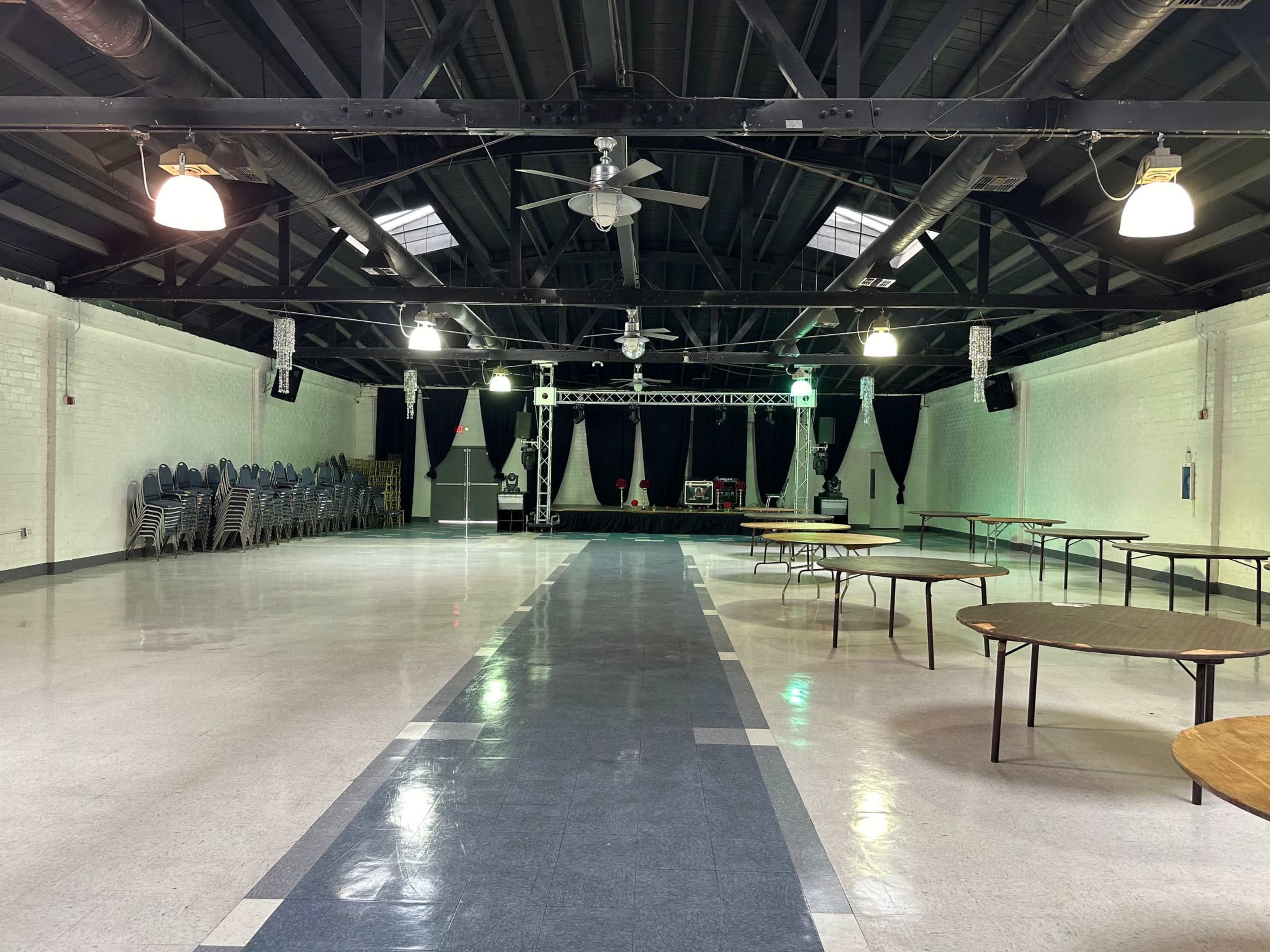 A large, vacant indoor hall features rows of stacked chairs on one side and several empty tables arranged throughout the space, with a stage at the far end.