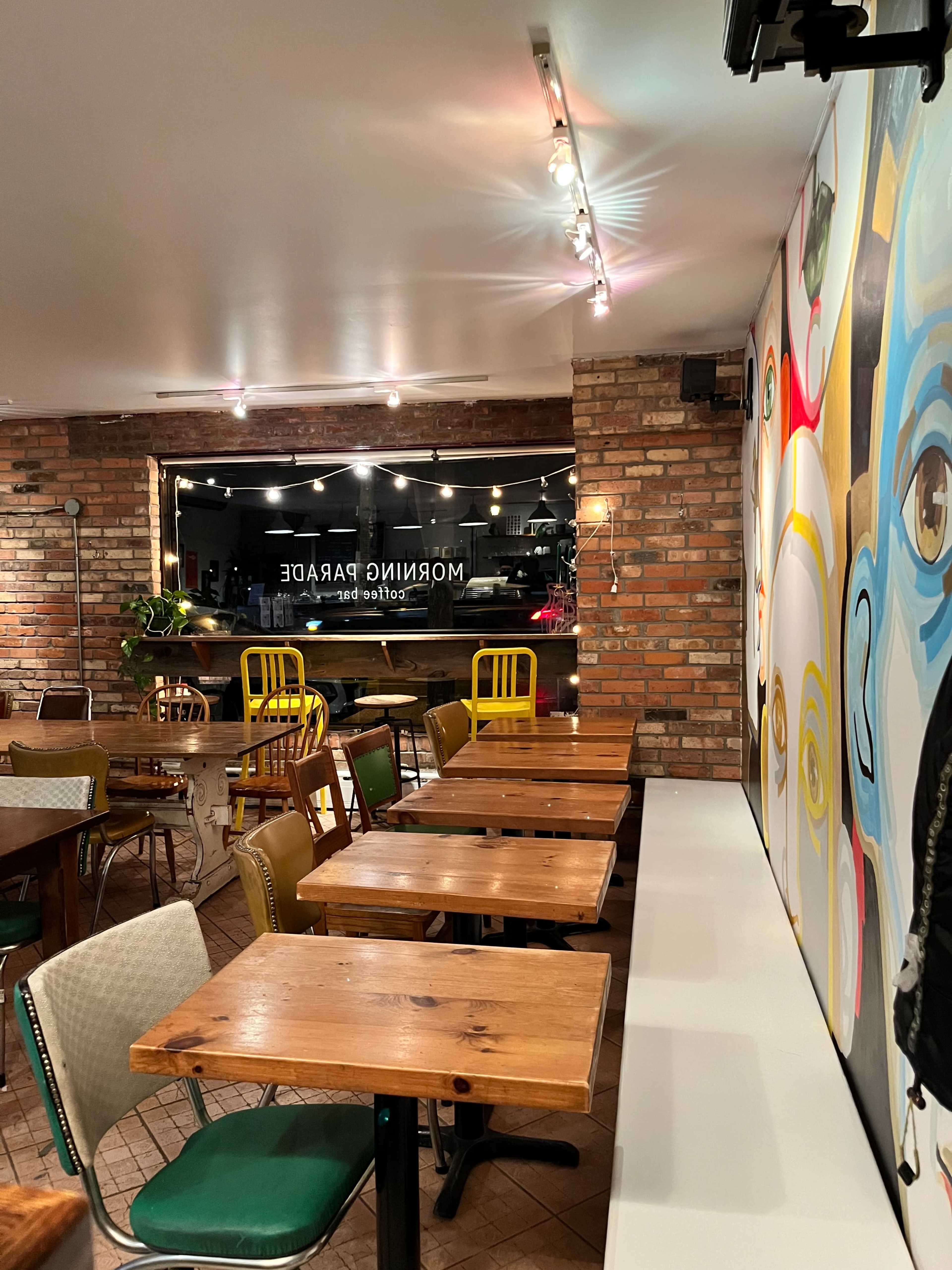 The image shows an interior view of a café with wooden tables, colorful chairs, and a mural on the wall.