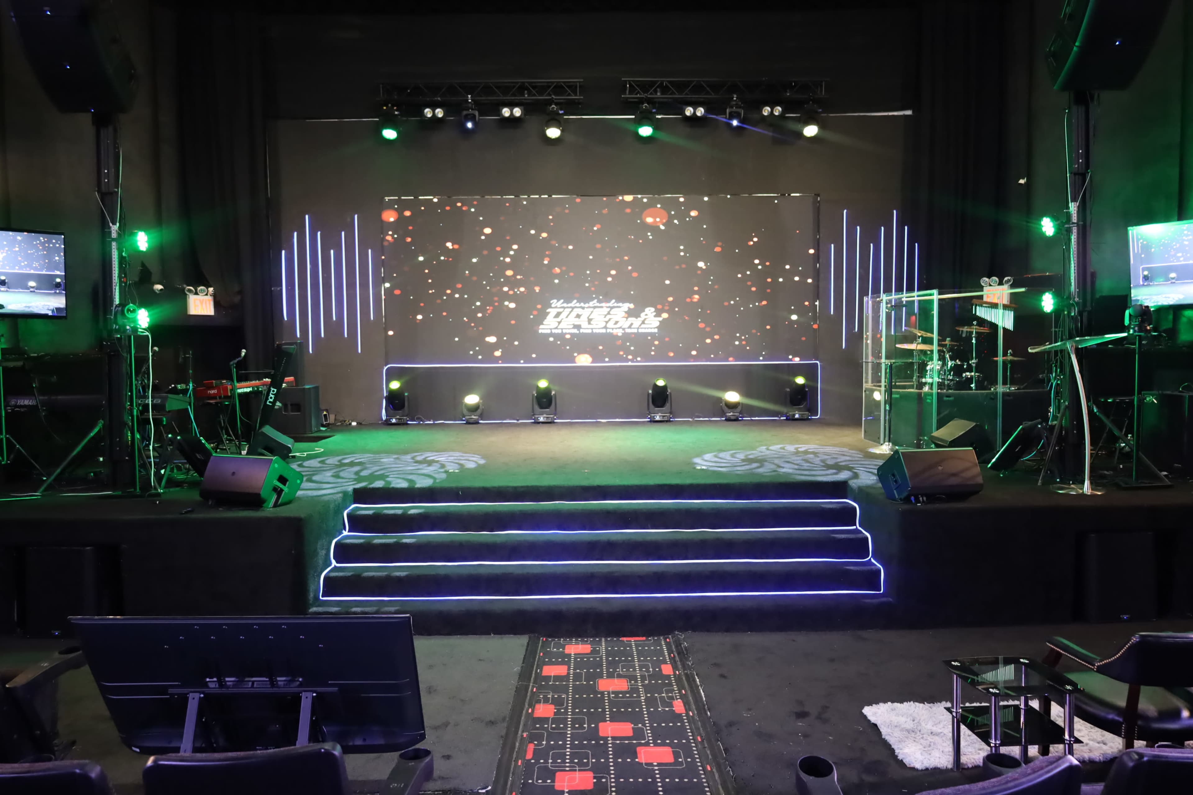 The image shows a stage setup with an elaborate lighting system, a large digital screen, and musical instruments arranged in a darkened venue.