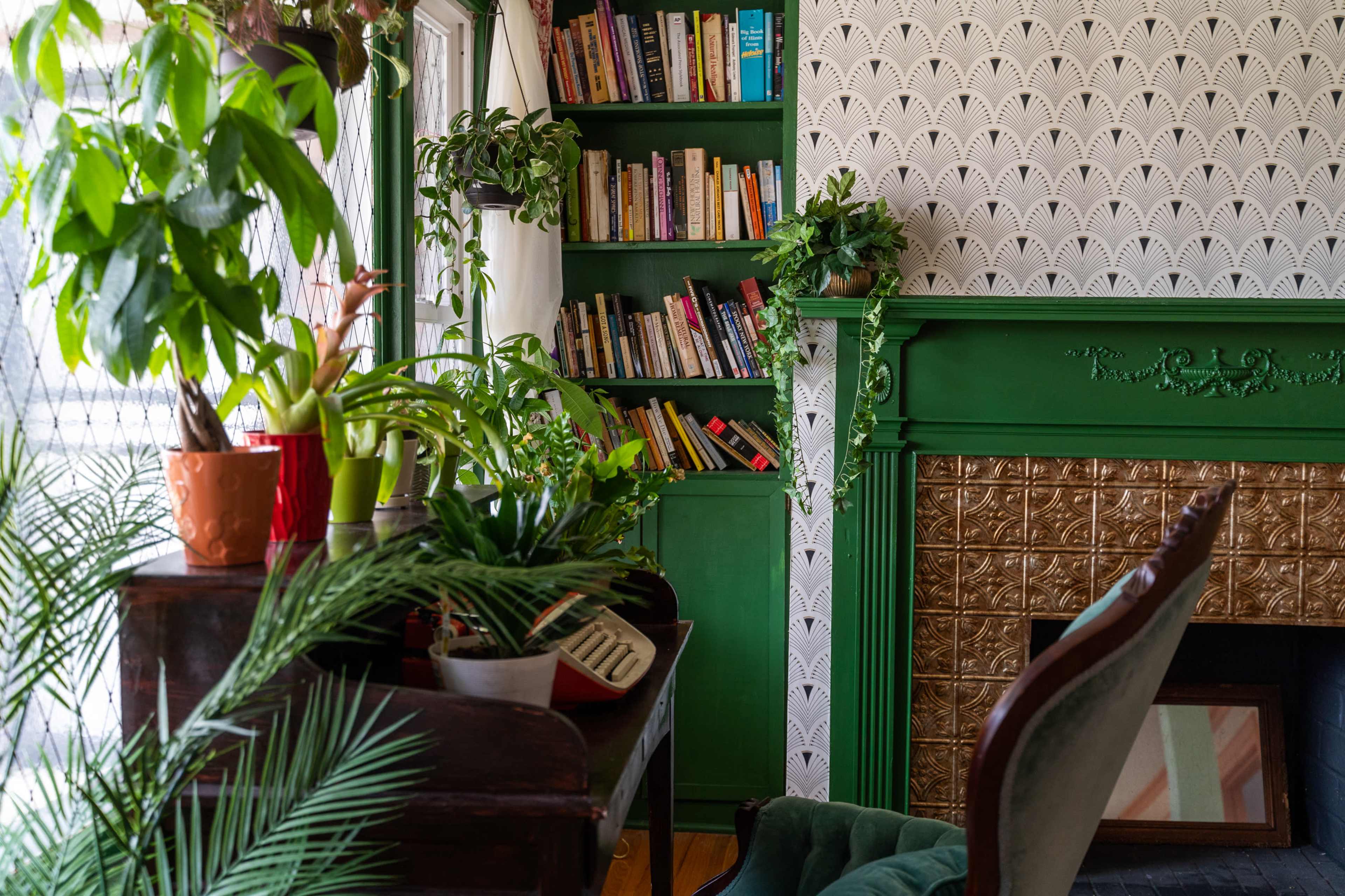 A cozy room features a green bookshelf filled with books, a decorative fireplace, and plants arranged on a wooden desk and shelves.