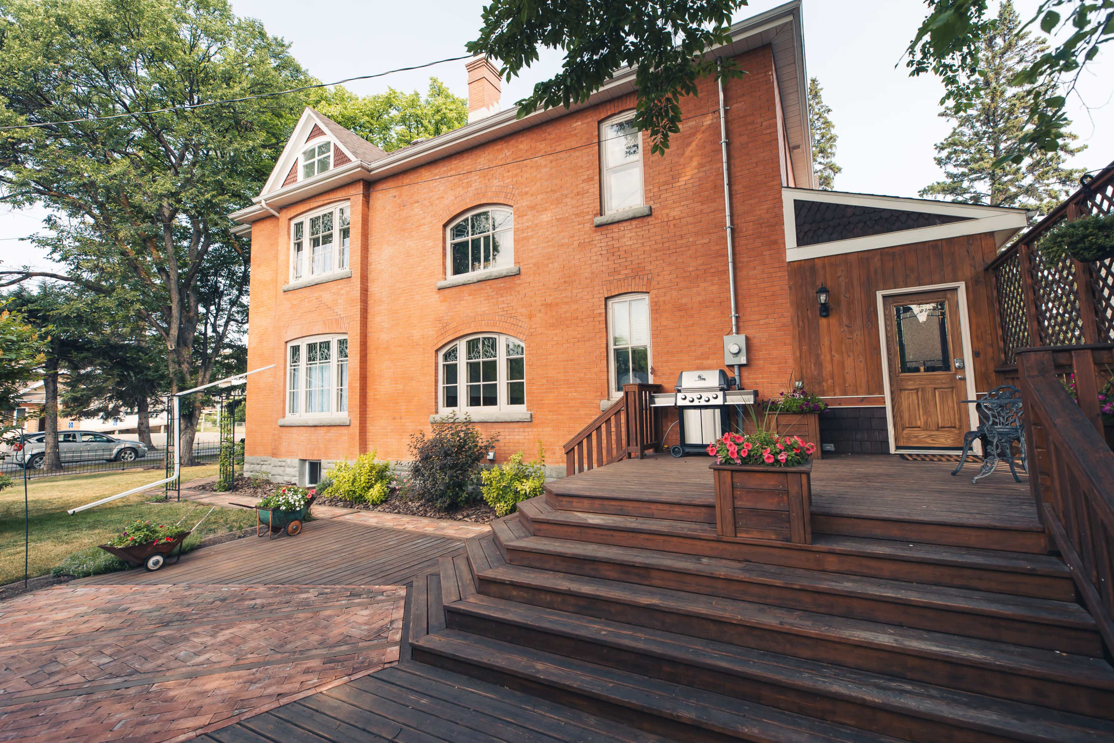 A two-story brick house with large windows and a wooden deck is surrounded by landscaped greenery and potted plants.