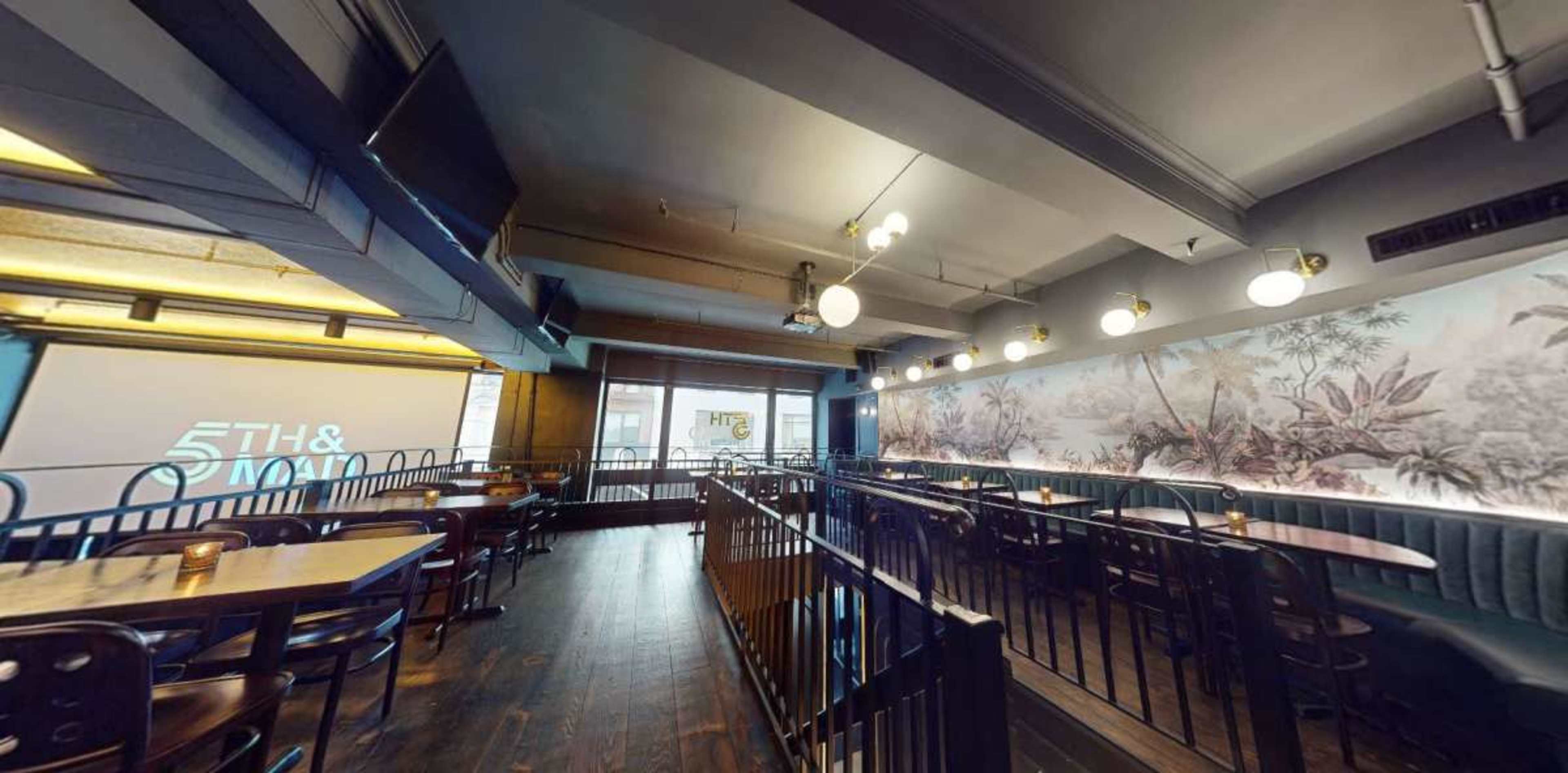 The image shows a modern restaurant interior featuring wooden tables, dark seating, and decorative wall art illuminated by overhead lighting.